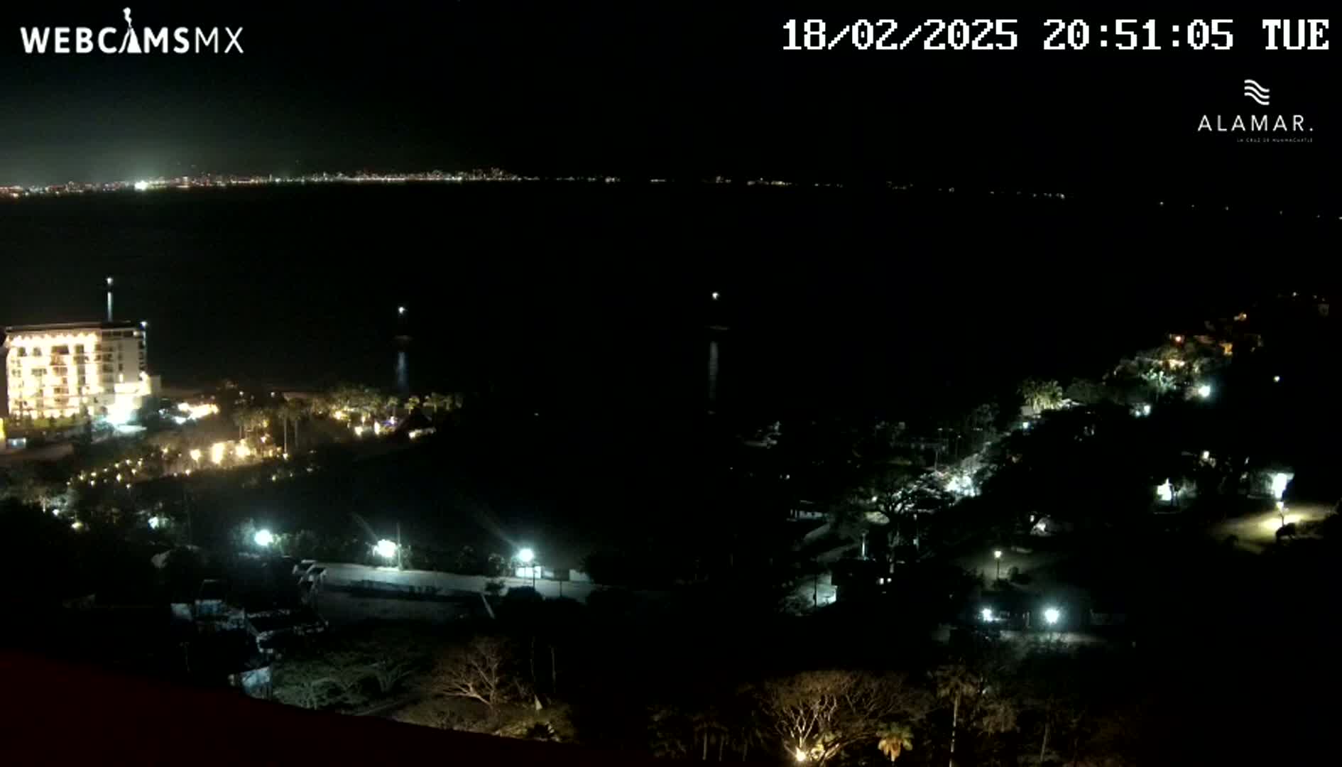 A nighttime, high-angle view shows a coastal area with a lit-up building near the water, other buildings and lights scattered across a dark landscape, and a city skyline faintly visible across the water in the distance.
