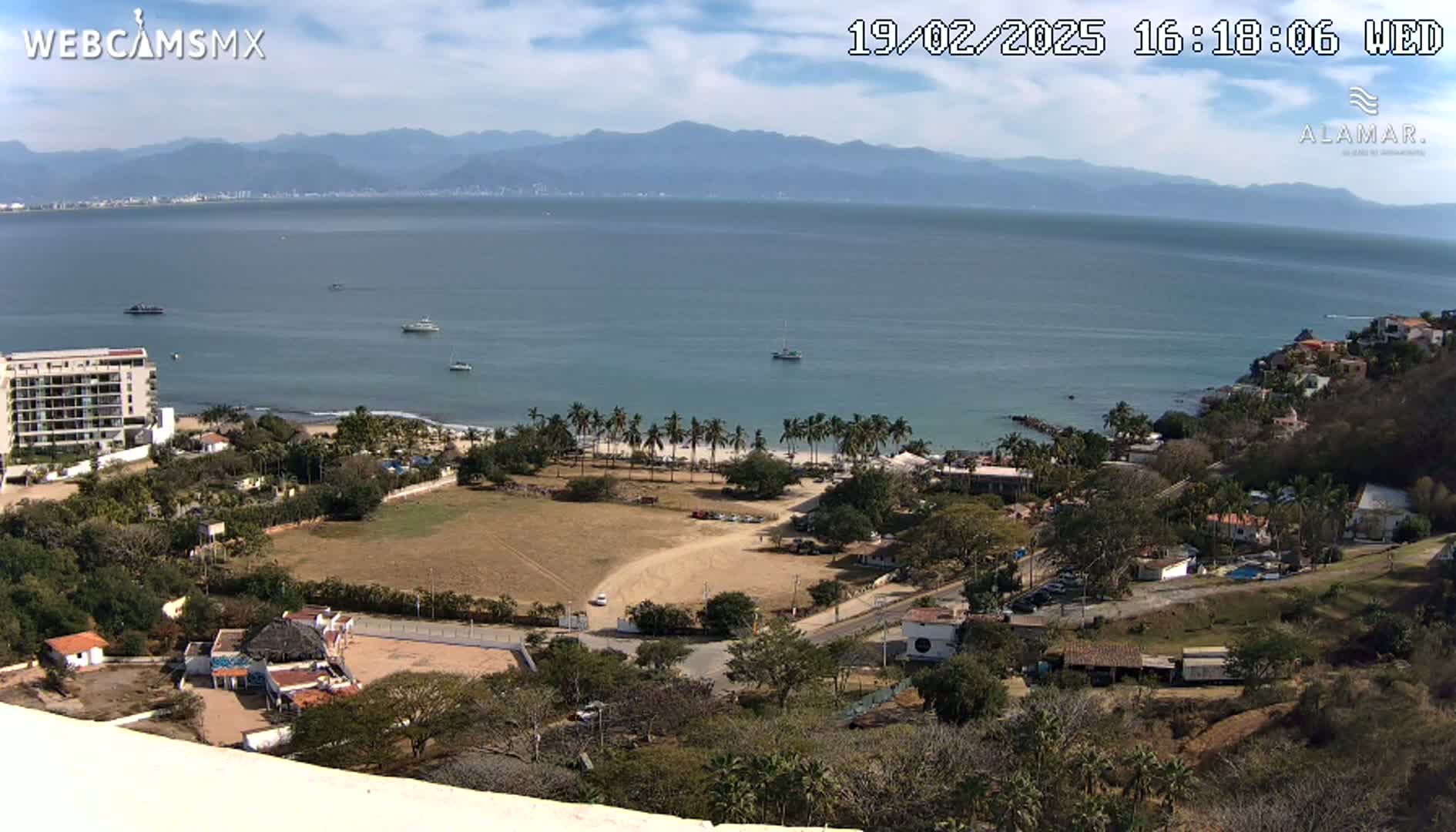 An elevated view of a coastal town on a partly sunny day, showing a beach with palm trees, a large body of water with boats, and mountains in the distance.