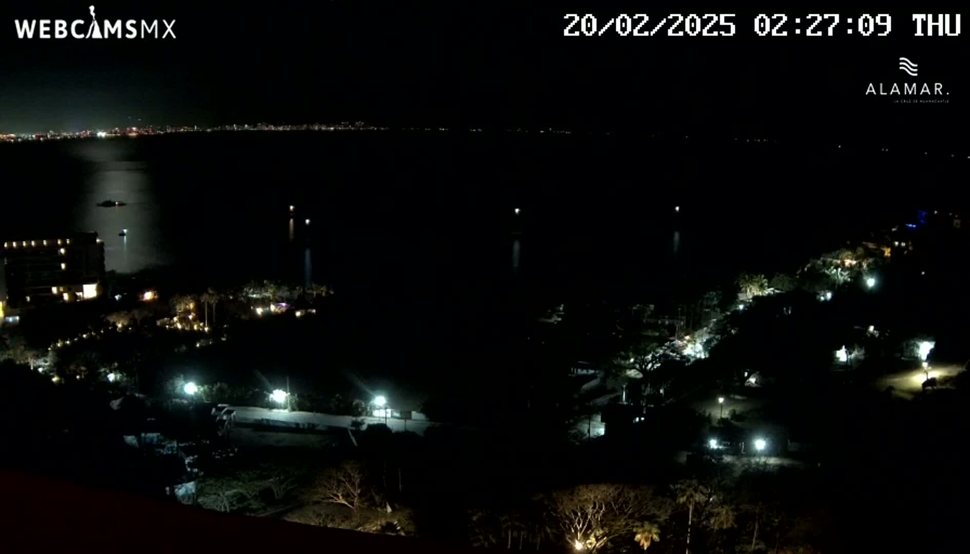 A nighttime, high-angle view shows a dark body of water with a distant city skyline, and a stretch of land with sparsely lit buildings and vegetation.