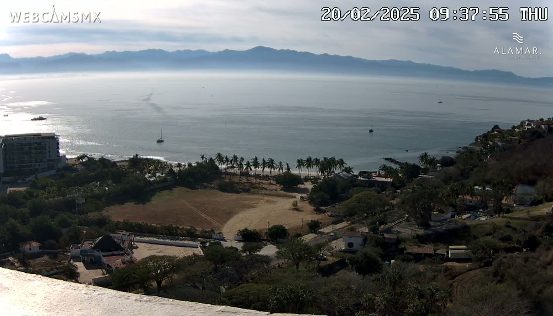 A high-angle, long shot shows a tranquil ocean scene under a mostly clear sky, with a beach, palm trees, buildings, and hills visible from an elevated vantage point.