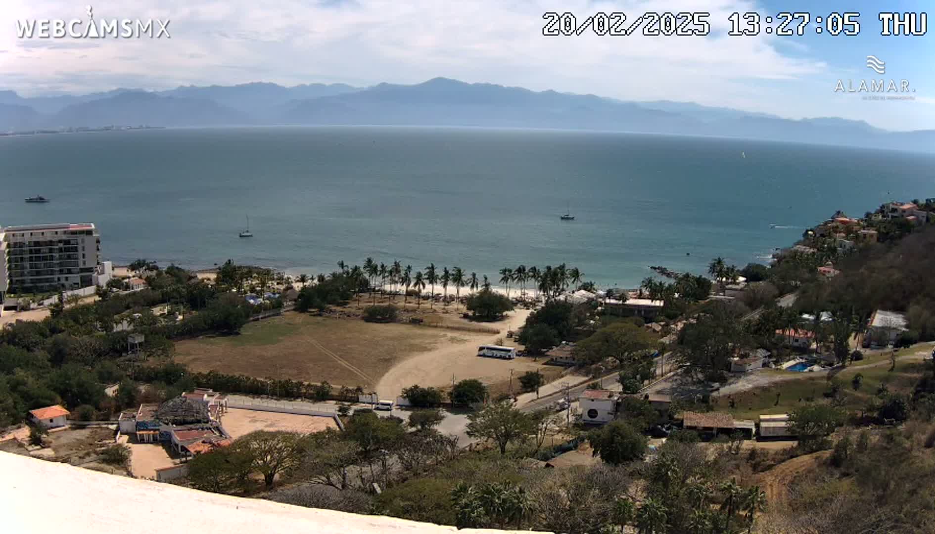 An elevated view shows a coastline with a beach, palm trees, buildings, and a calm ocean under a mostly clear sky.