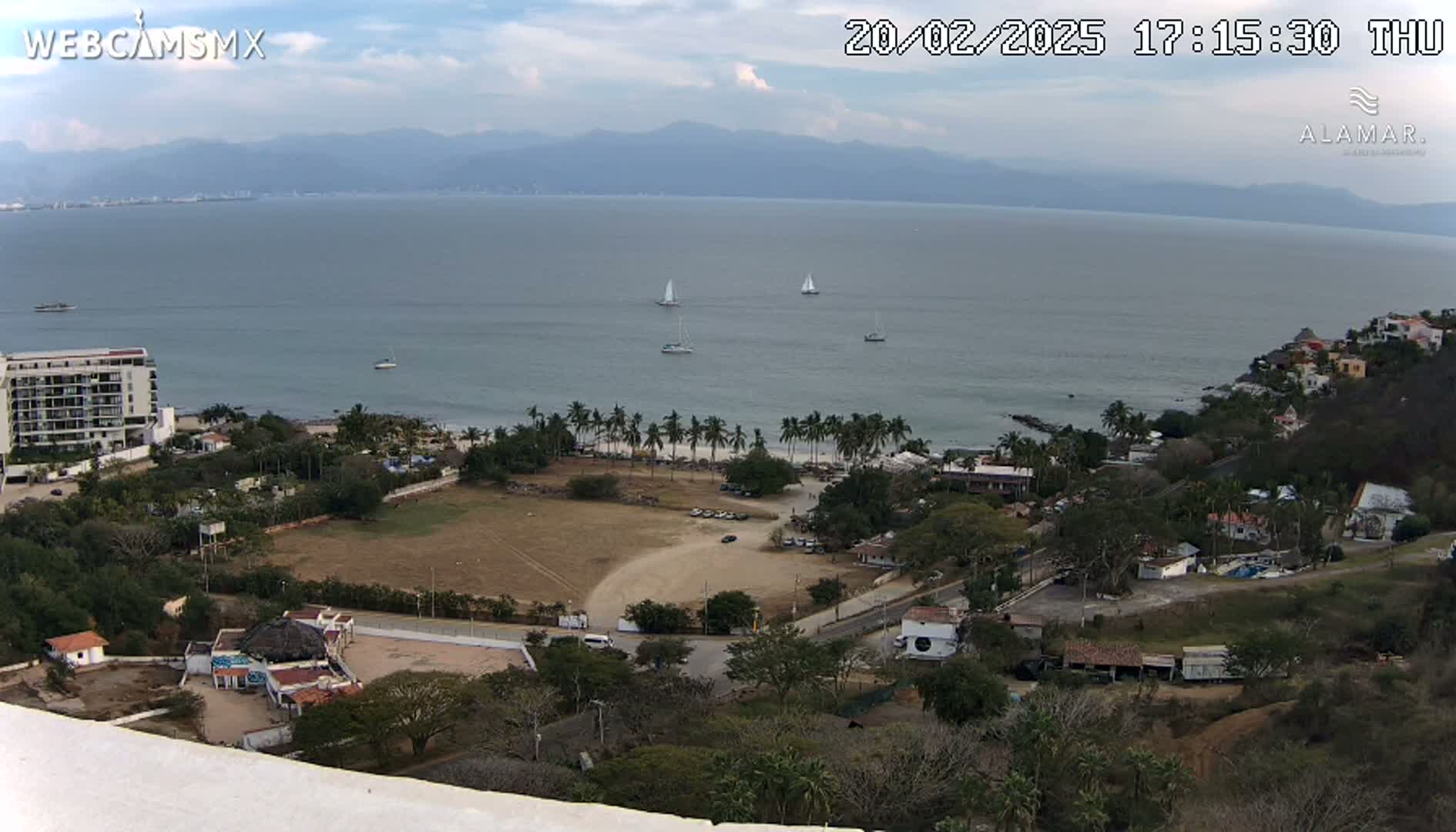 An elevated view shows a coastal town on a partly cloudy day, with several sailboats visible on the calm ocean, mountains in the background, and a large open field next to the beach.