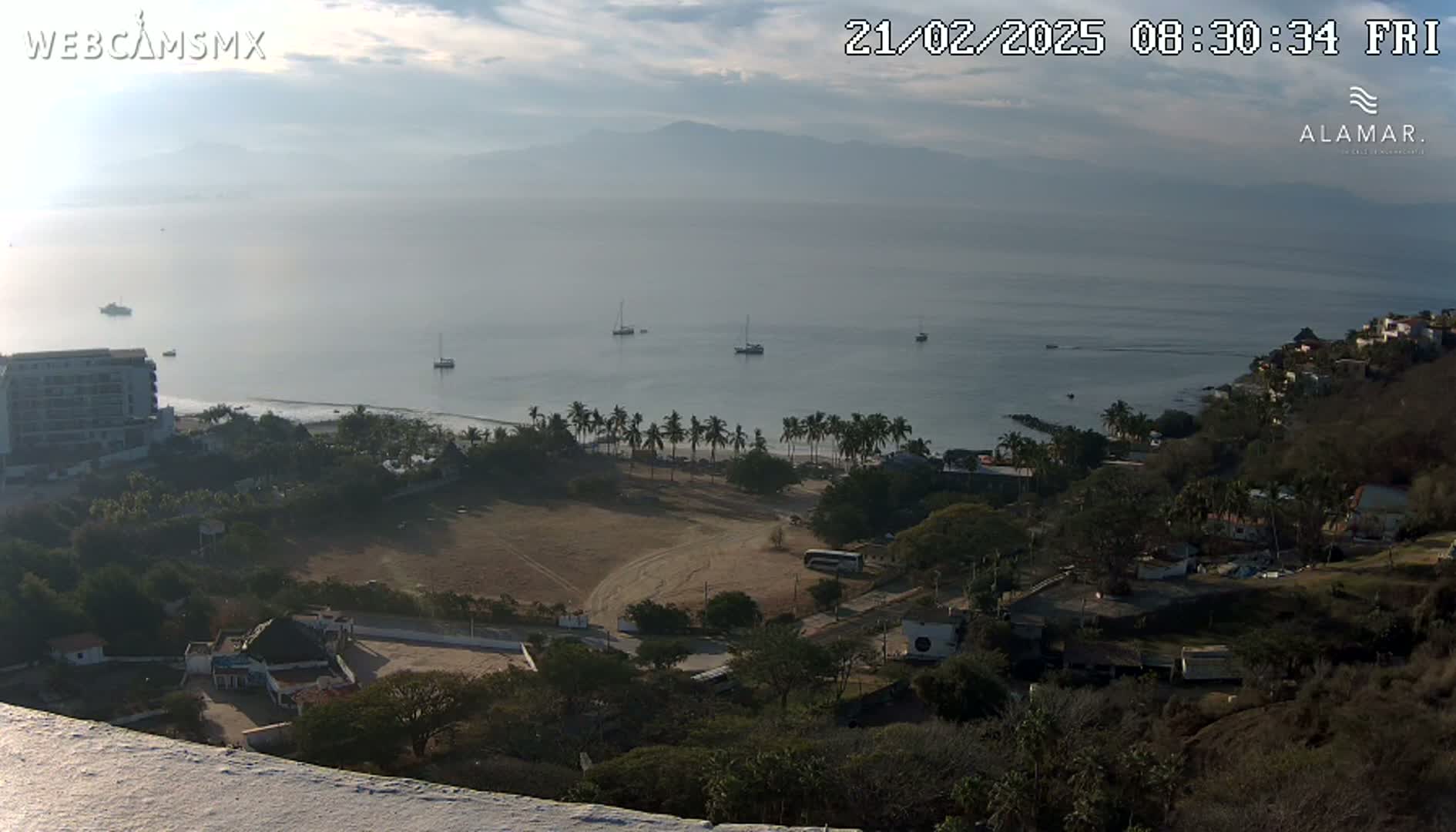 A hazy coastal scene shows a calm bay with several boats, a beach lined with palm trees, and a dry, open field, all viewed from an elevated position overlooking a hillside town.