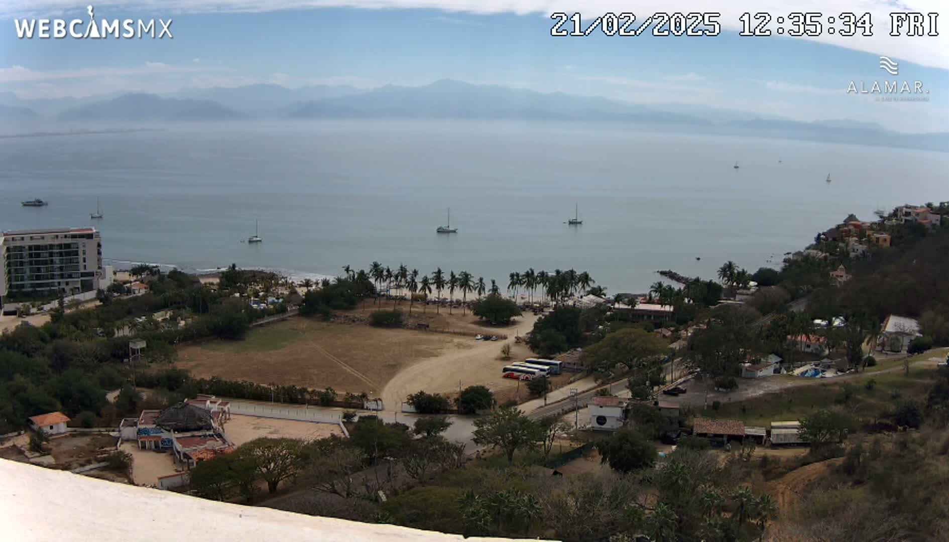 An elevated view of a coastal town on a partly cloudy day, showcasing a calm ocean with several boats, a sandy beach lined with palm trees, buildings, and a large empty field.