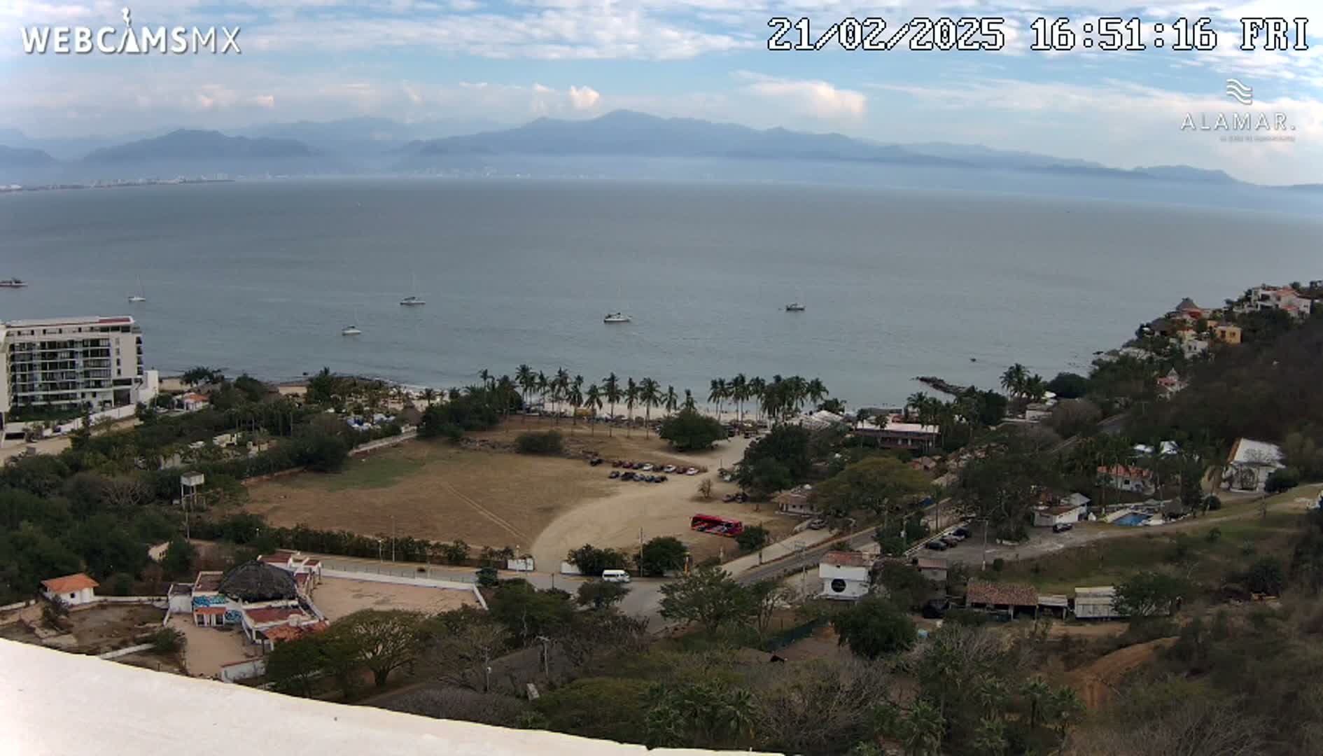 An elevated view shows a coastline with a calm ocean, a sandy beach lined with palm trees, buildings, and a parking lot, under a mostly clear sky.