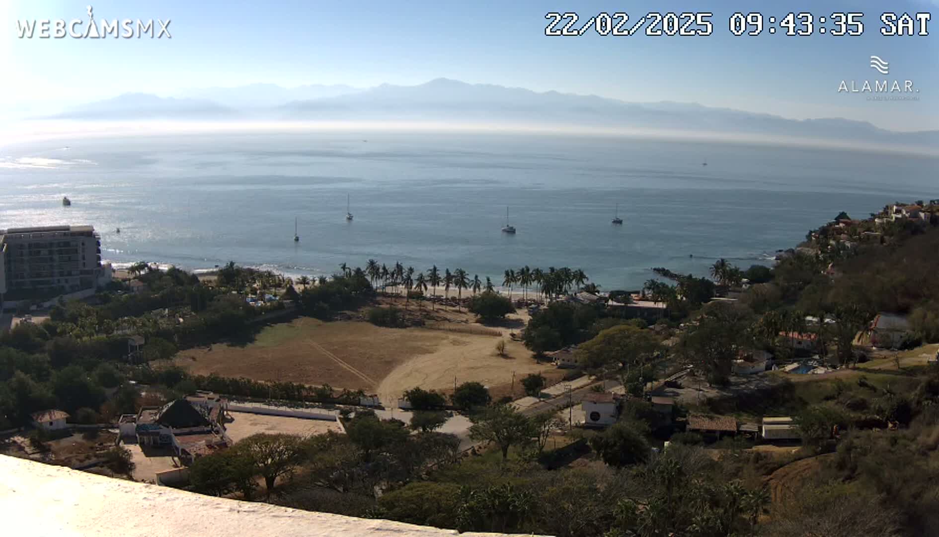 A high-angle, long shot on a clear day shows a coastline with a calm ocean, a sandy beach lined with palm trees, buildings nestled amongst vegetation on a hillside, and distant mountains under a clear sky.