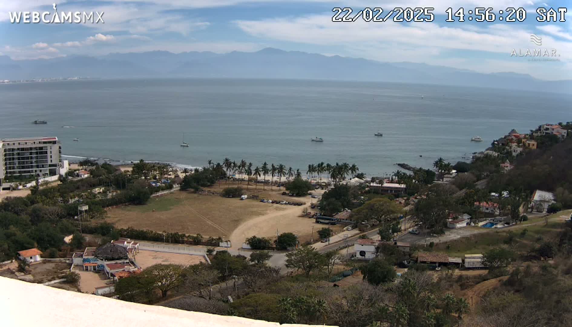 A high-angle view shows a coastal town on a sunny day, with a calm ocean and distant mountains under a partly cloudy sky.