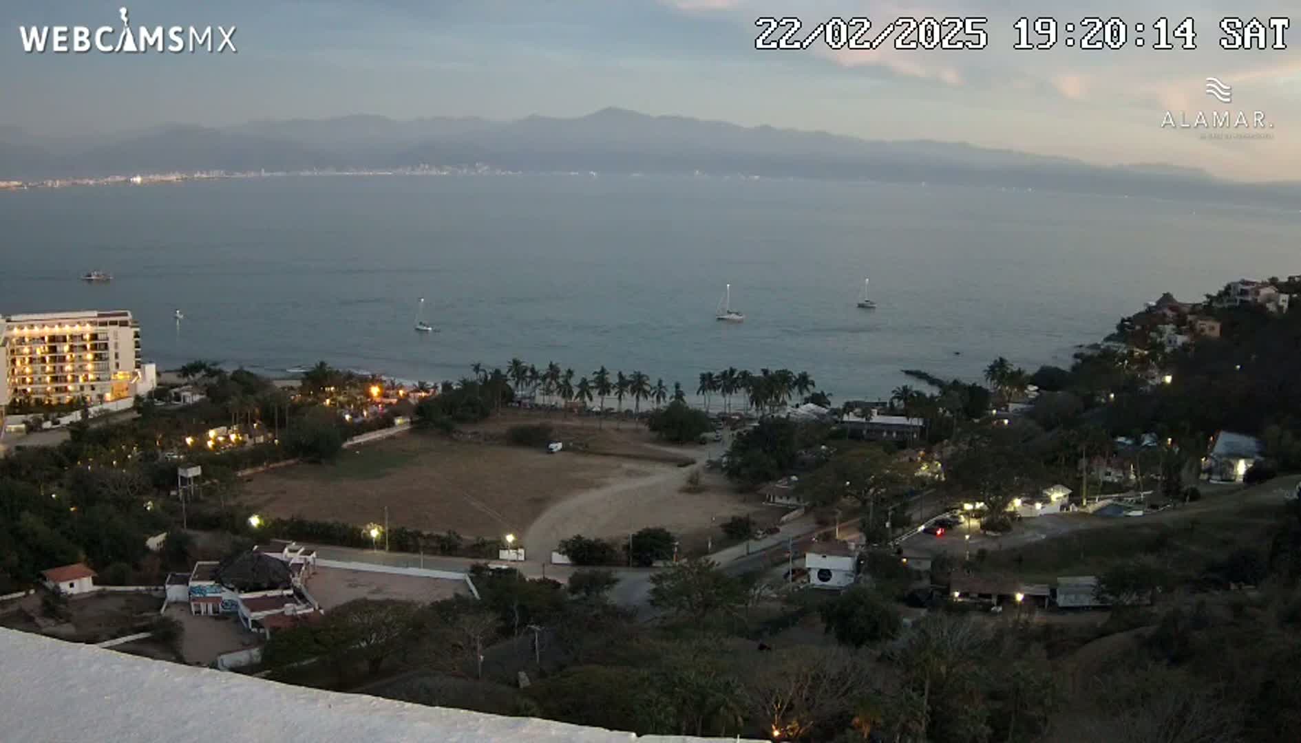 An elevated evening view shows a calm bay with several sailboats, a coastline with palm trees and buildings, and mountains in the distance under a twilight sky.