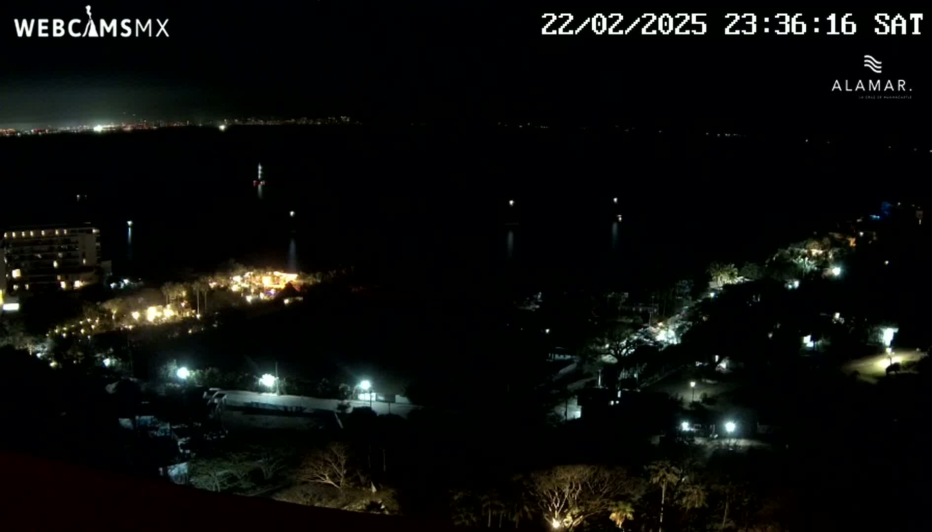 A nighttime, high-angle view shows a dark body of water with a few lights visible in the distance, a shoreline with sparsely lit buildings and vegetation, and a city skyline faintly visible in the far background.