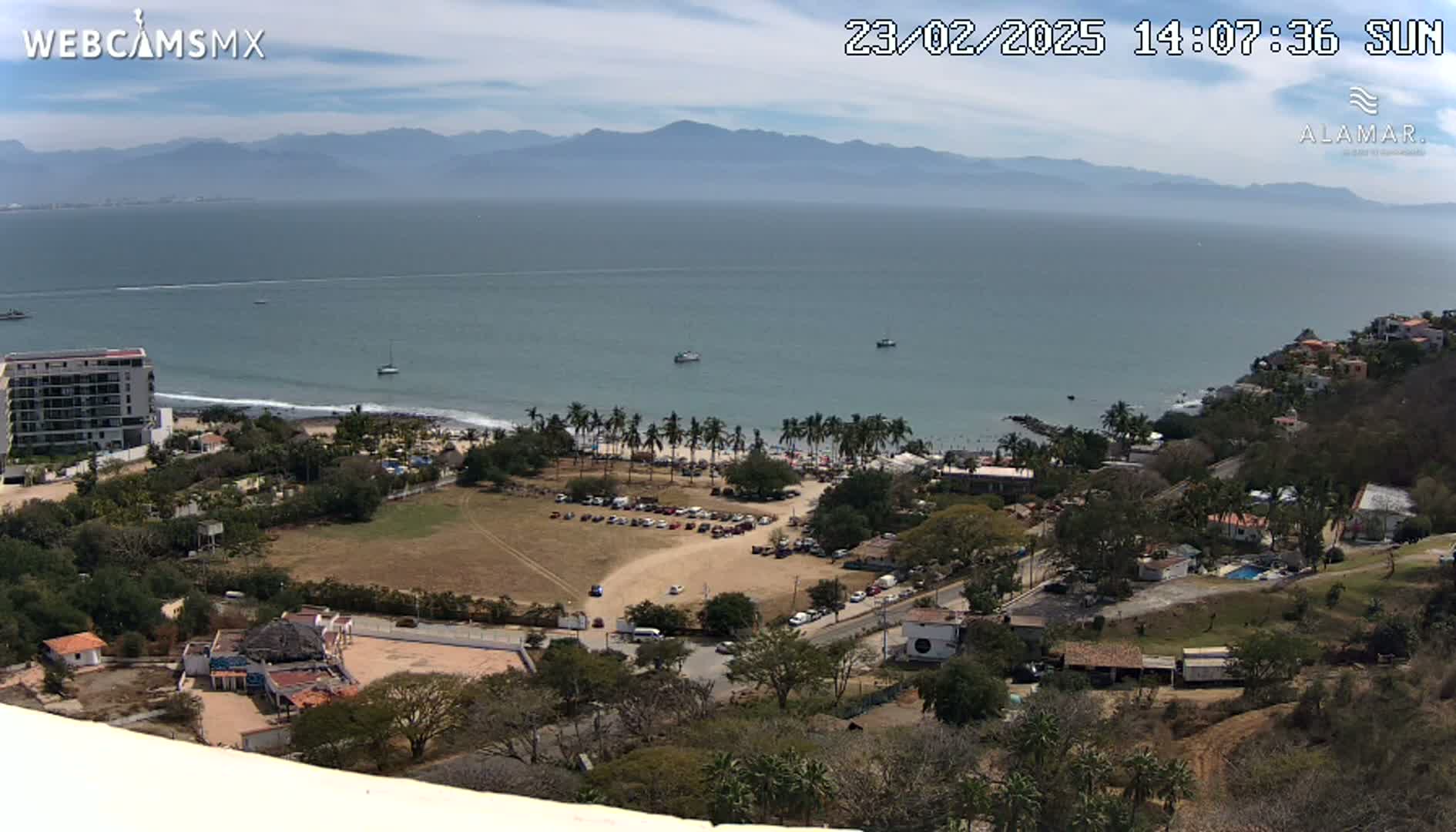 A sunny, high-angle view shows a coastline with a beach, palm trees, buildings, and a parking lot, all overlooking a calm ocean and distant mountains.