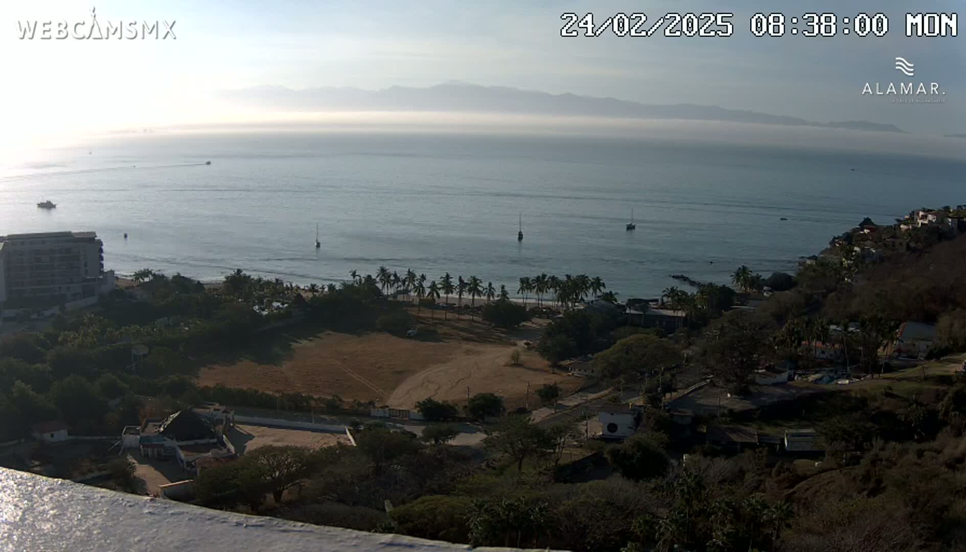 A high-angle, long shot of a coastal scene on a hazy morning shows a calm ocean with several boats, a sandy beach lined with palm trees, and a hillside town with buildings and vegetation.