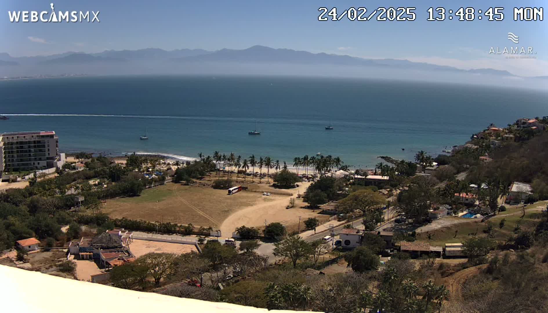 An elevated view shows a coastal town on a sunny day, with a calm ocean stretching to distant mountains.