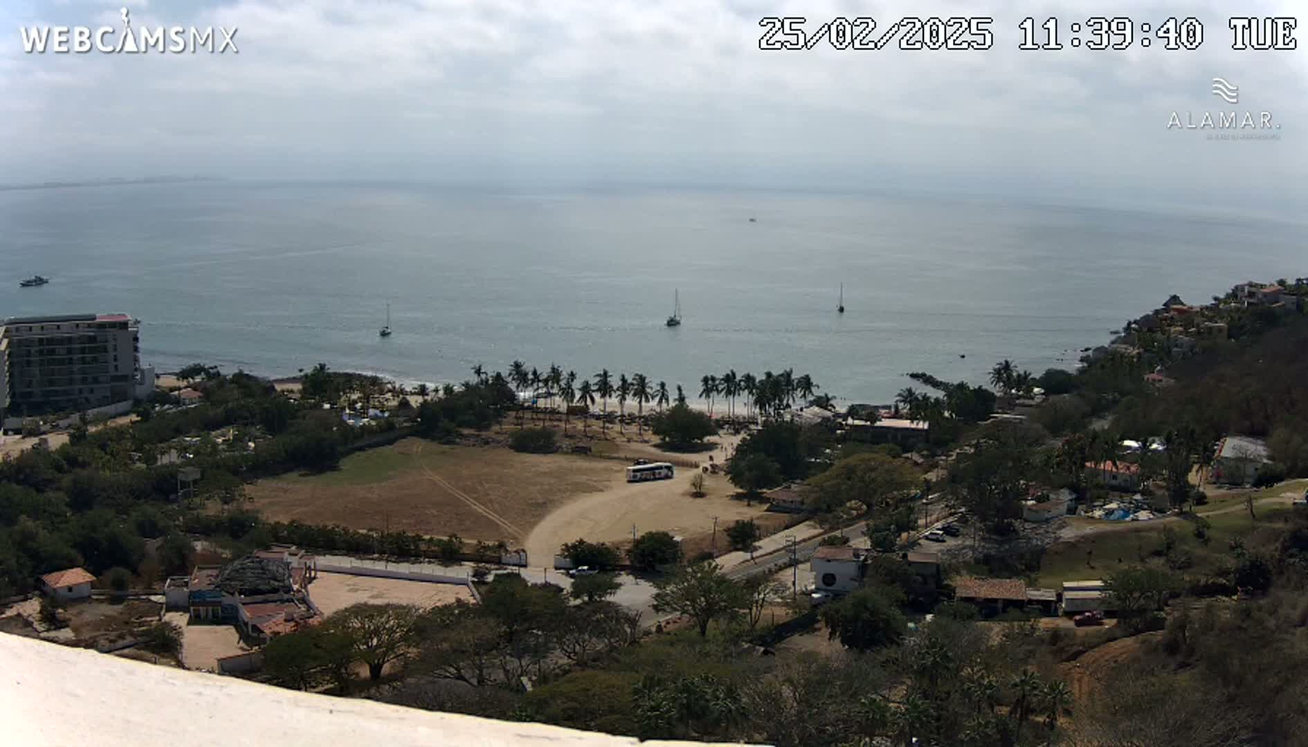 An elevated view of a coastal town on a partly cloudy day, showing a calm ocean with a few boats, a sandy beach with palm trees, and buildings nestled amongst vegetation.