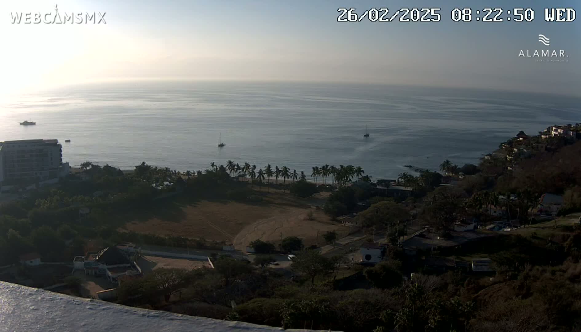 A high-angle, long shot shows a calm ocean under a hazy sunrise, with a coastline featuring buildings, palm trees, and sparsely vegetated land.