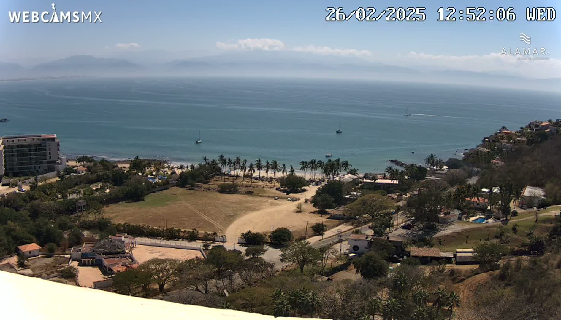 A high-angle view shows a coastal town on a sunny day, with a sandy beach lined with palm trees and a calm ocean extending to distant mountains.