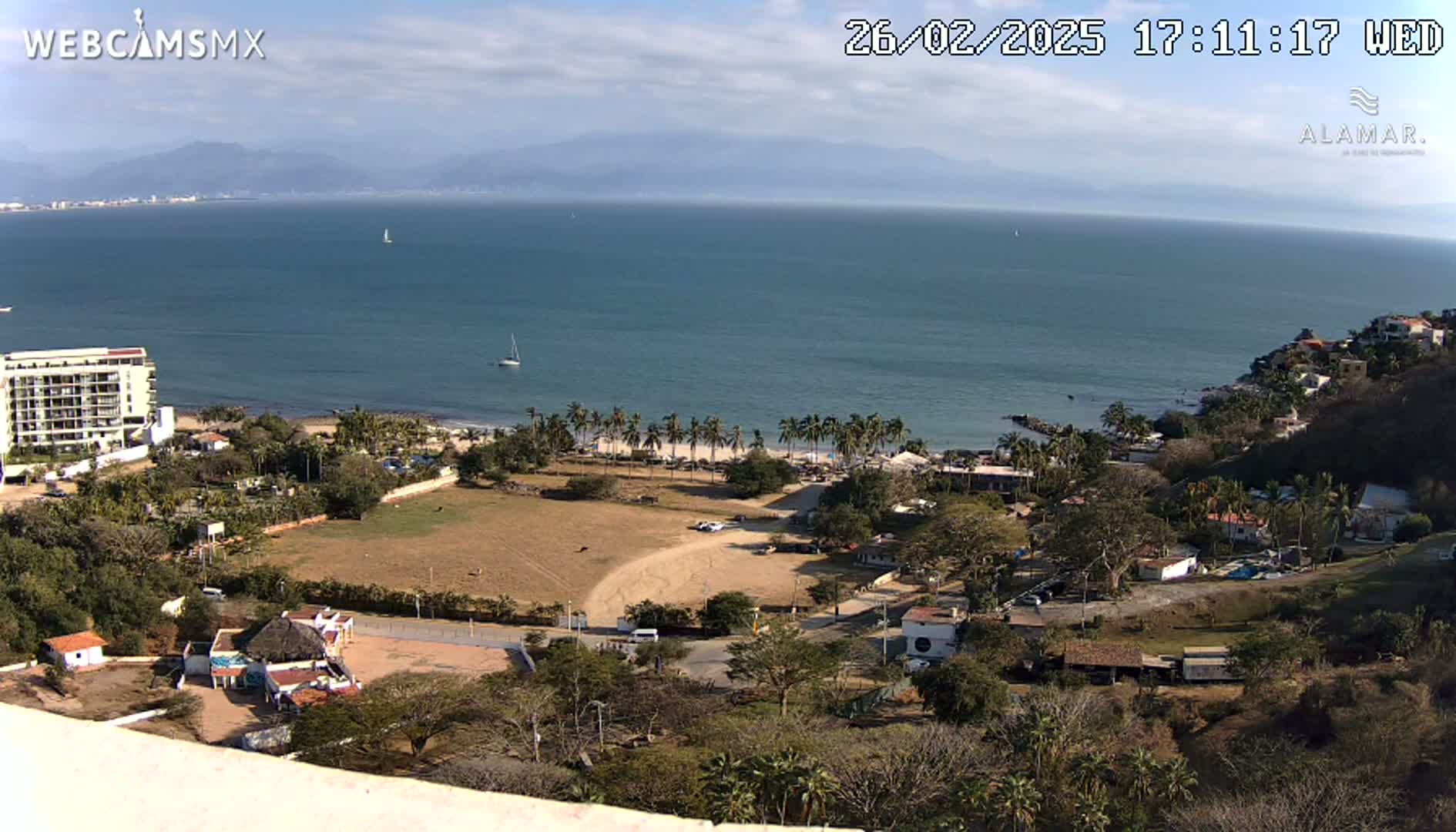 A coastal scene on a partly cloudy day shows a beach with palm trees, a building complex, and a large expanse of calm ocean with mountains in the distance.