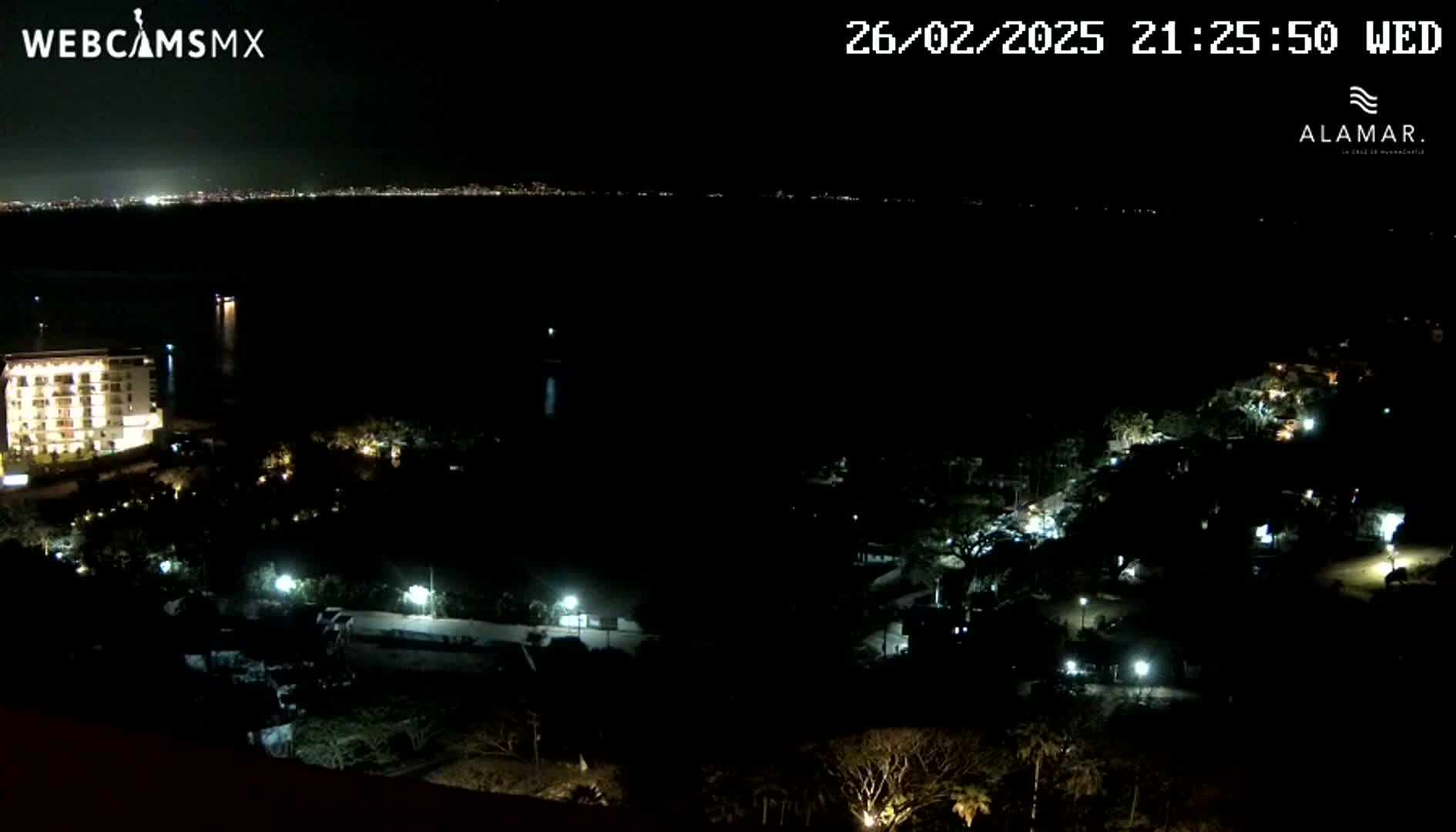 A nighttime view of a city coastline, showing a dark body of water, illuminated buildings, and sparsely lit vegetation.