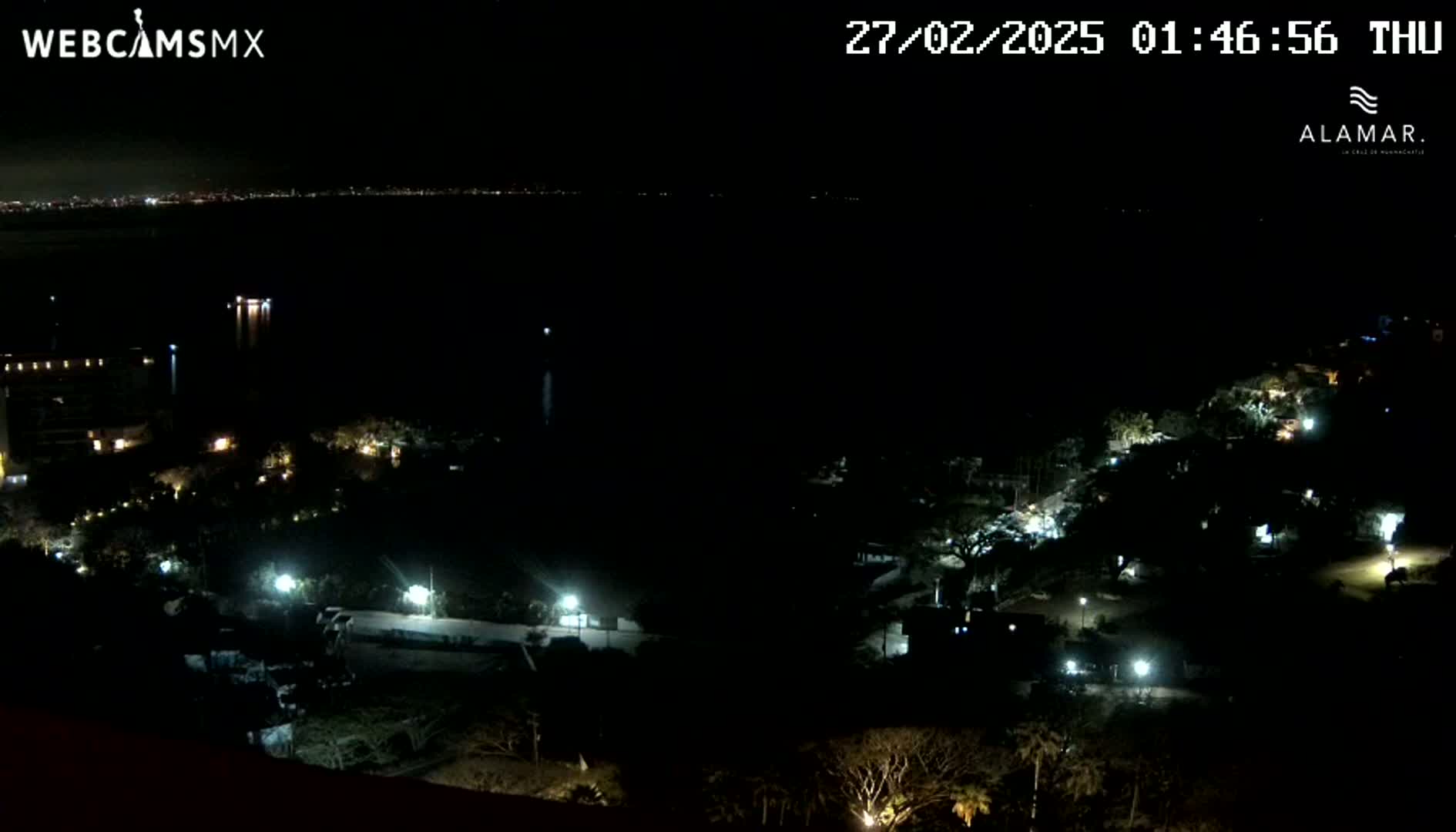 A nighttime, high-angle view shows a dark body of water with a city skyline in the distance and sparsely lit buildings and vegetation on the foreground.