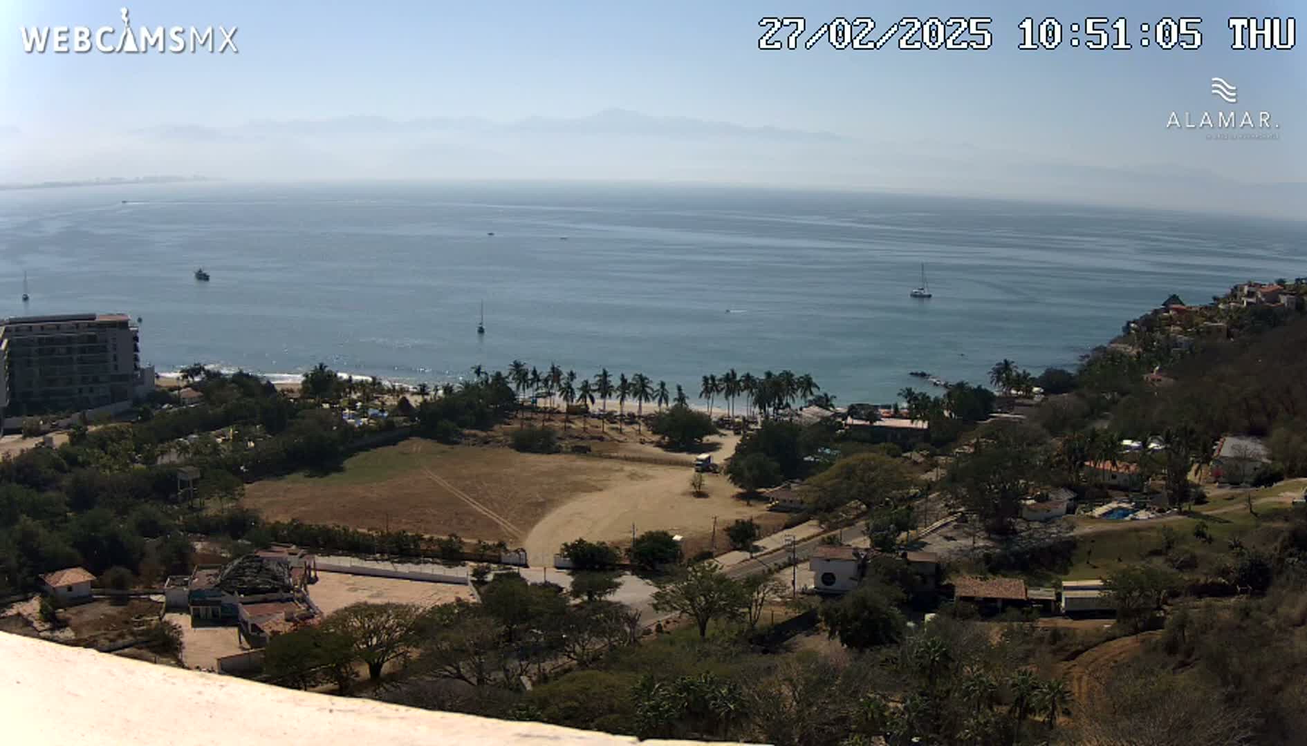 A high-angle, long shot shows a coastal scene on a sunny day, with a calm ocean, a sandy beach lined with palm trees, and a hillside dotted with buildings and vegetation.
