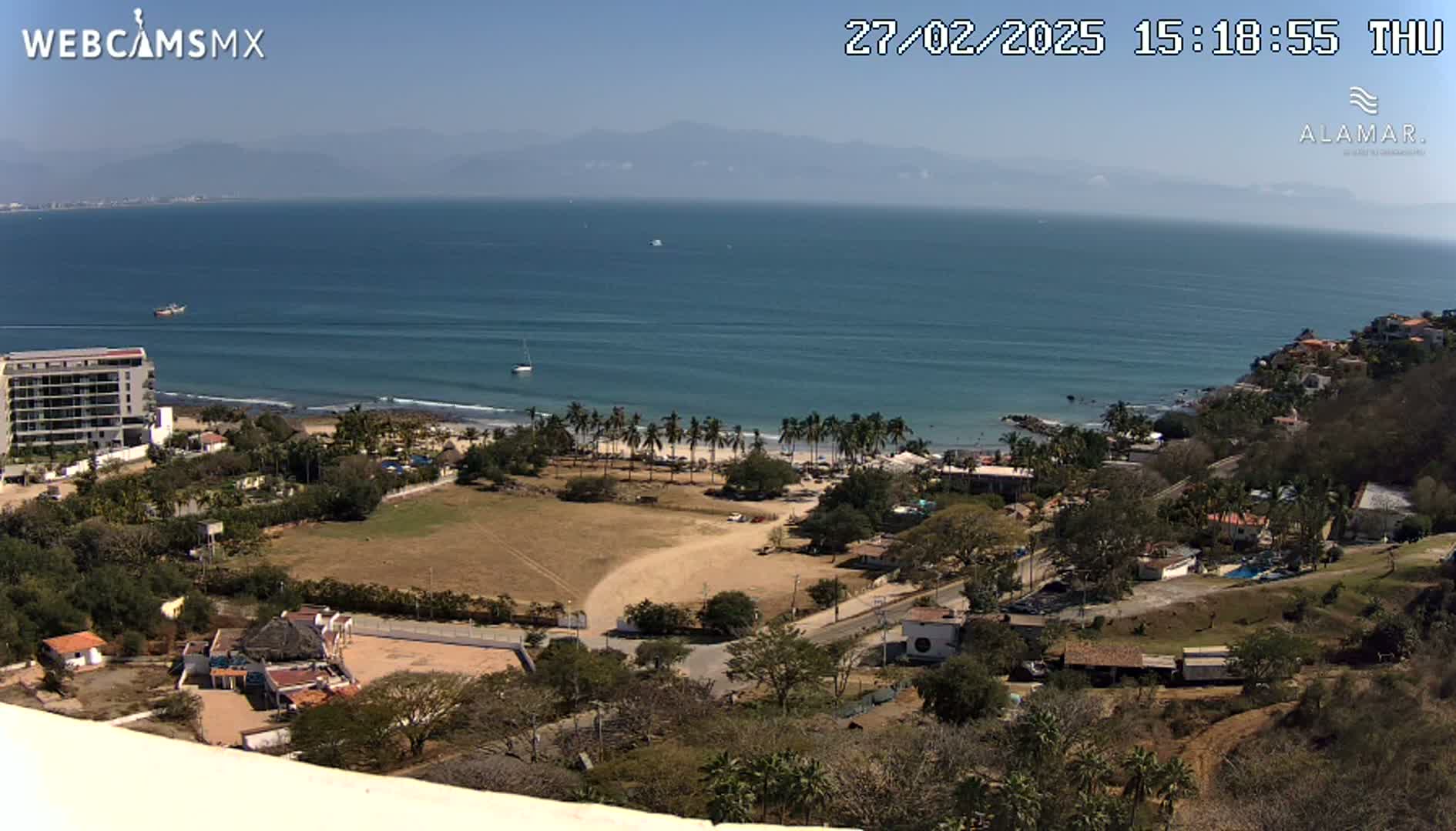 An elevated view of a coastline on a sunny day shows a calm ocean with a sandy beach lined with palm trees, buildings, and vegetation on the hillside.
