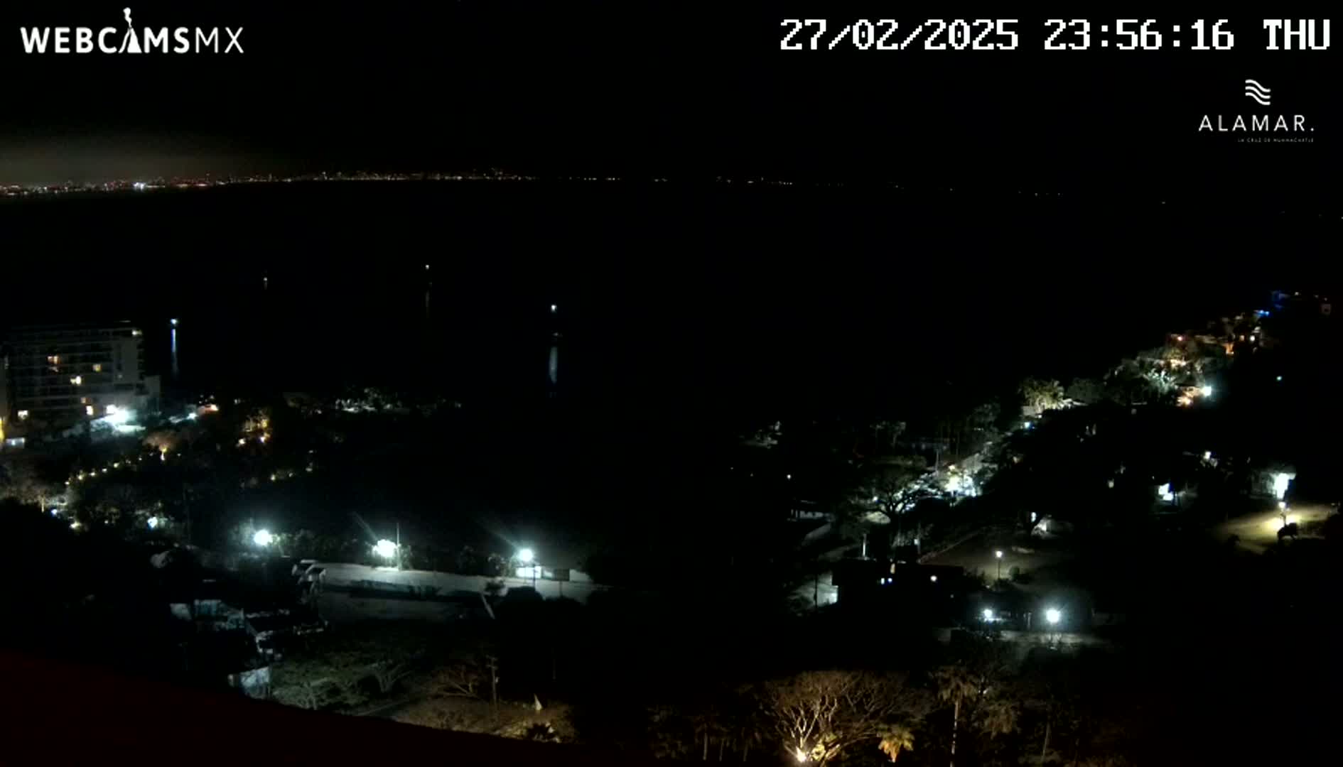 A nighttime, high-angle view shows a dark coastline with scattered lights from buildings and streetlights, overlooking a body of dark water with a distant city skyline faintly visible.