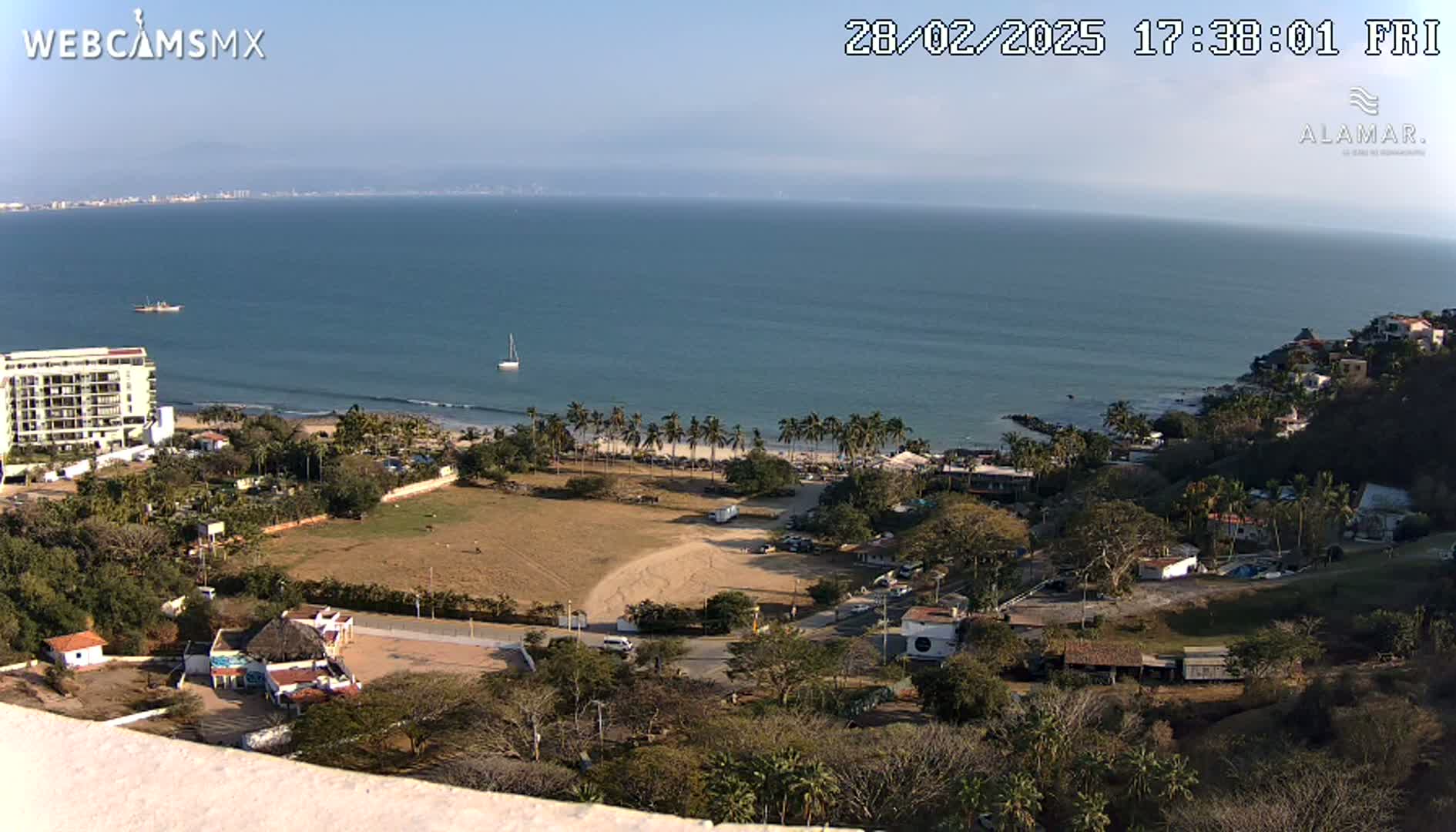 A high-angle view shows a calm ocean with a sandy beach lined with palm trees, overlooking a town nestled in hills under a clear, sunny sky.