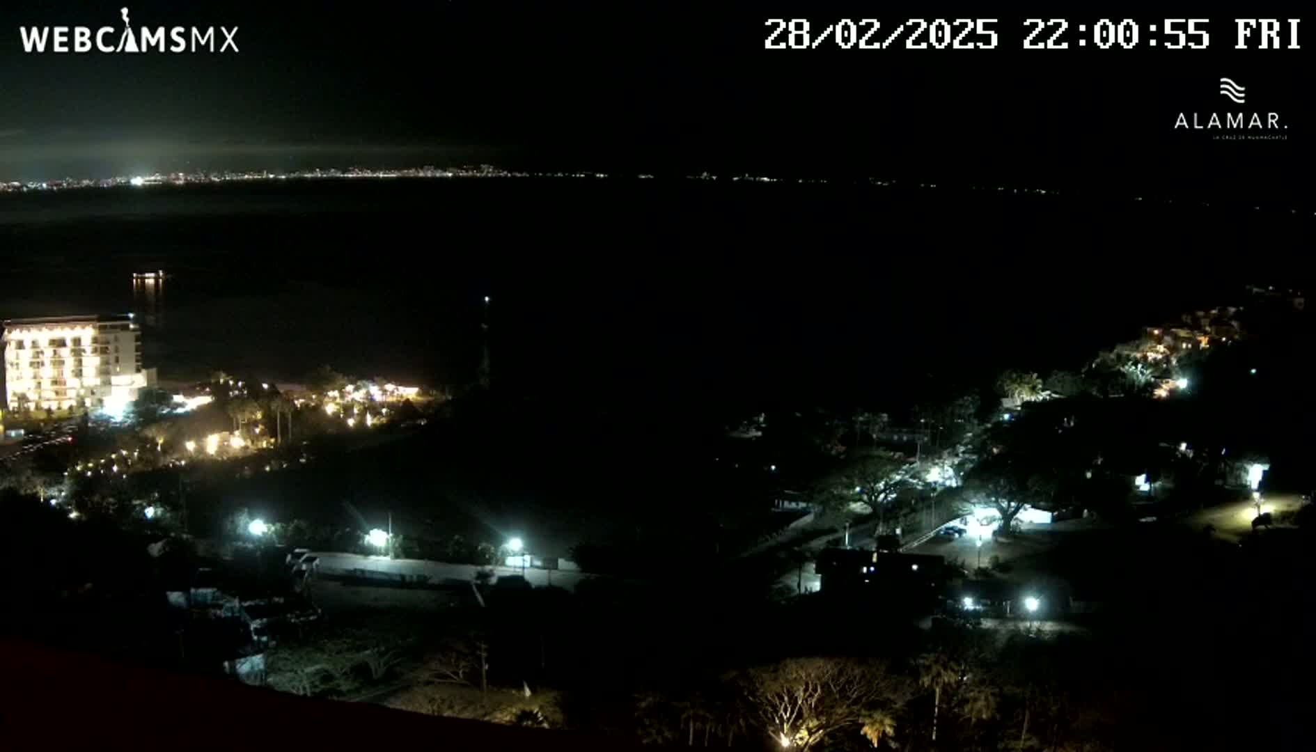 A nighttime coastal scene shows a dark body of water with a city skyline faintly visible in the distance, and a dark, sparsely lit area of land closer to the viewer with a few buildings and lights.
