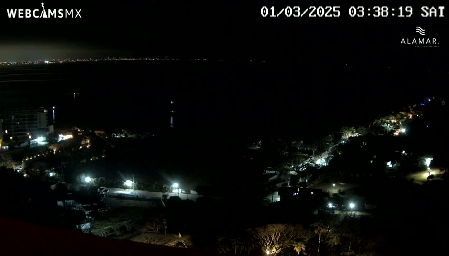 A nighttime, high-angle view shows a dark body of water with a distant city skyline, and a coastal area with sparsely lit buildings and vegetation.