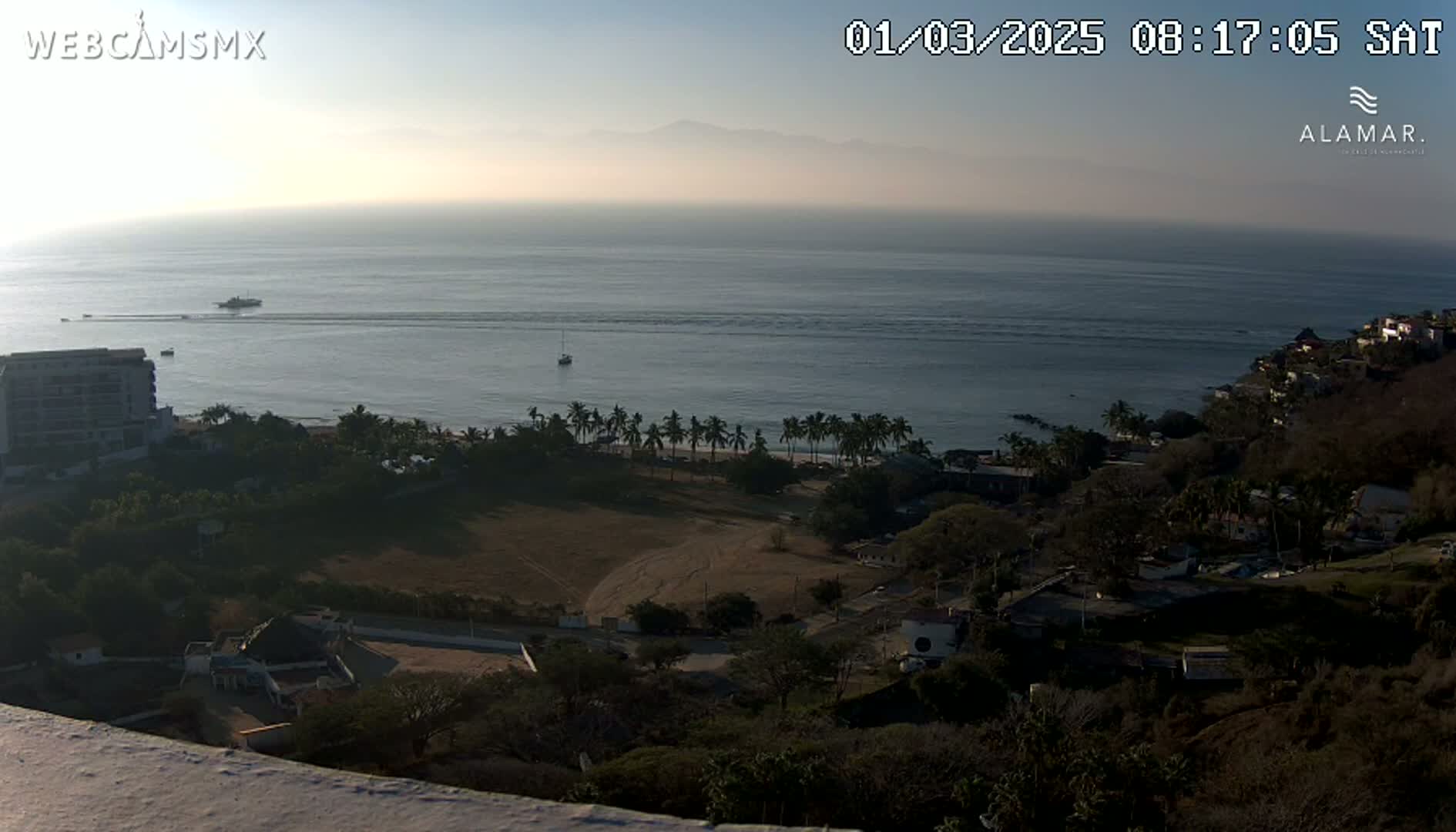 A hazy sunrise reveals a coastal scene with a calm ocean, a beach lined with palm trees, and buildings nestled amongst the hills.