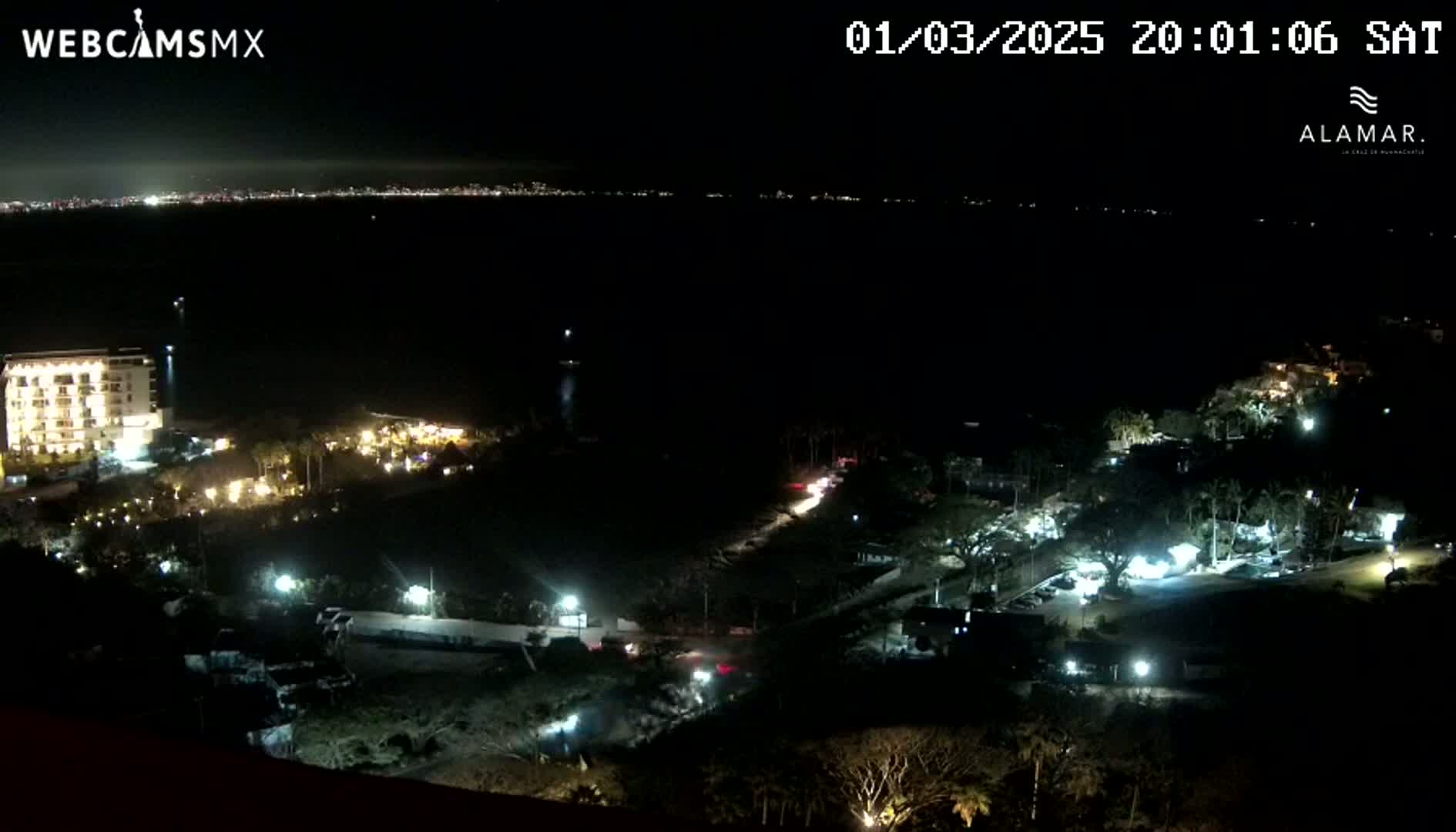 A nighttime coastal scene shows a dark body of water with a distant city skyline, illuminated buildings and vegetation closer to the viewer, and a few vehicles traveling on a road.
