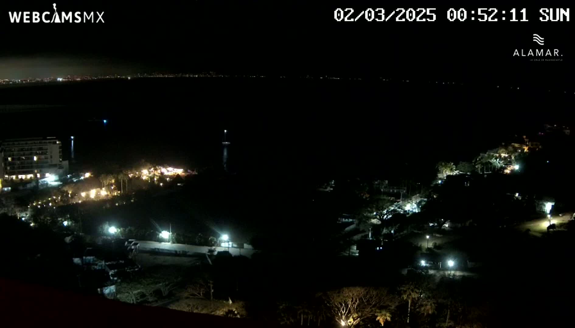 A nighttime view of a coastal area shows a dark body of water, a brightly lit building, and sparsely lit vegetation leading down to the water's edge, with a distant city skyline faintly visible in the background.