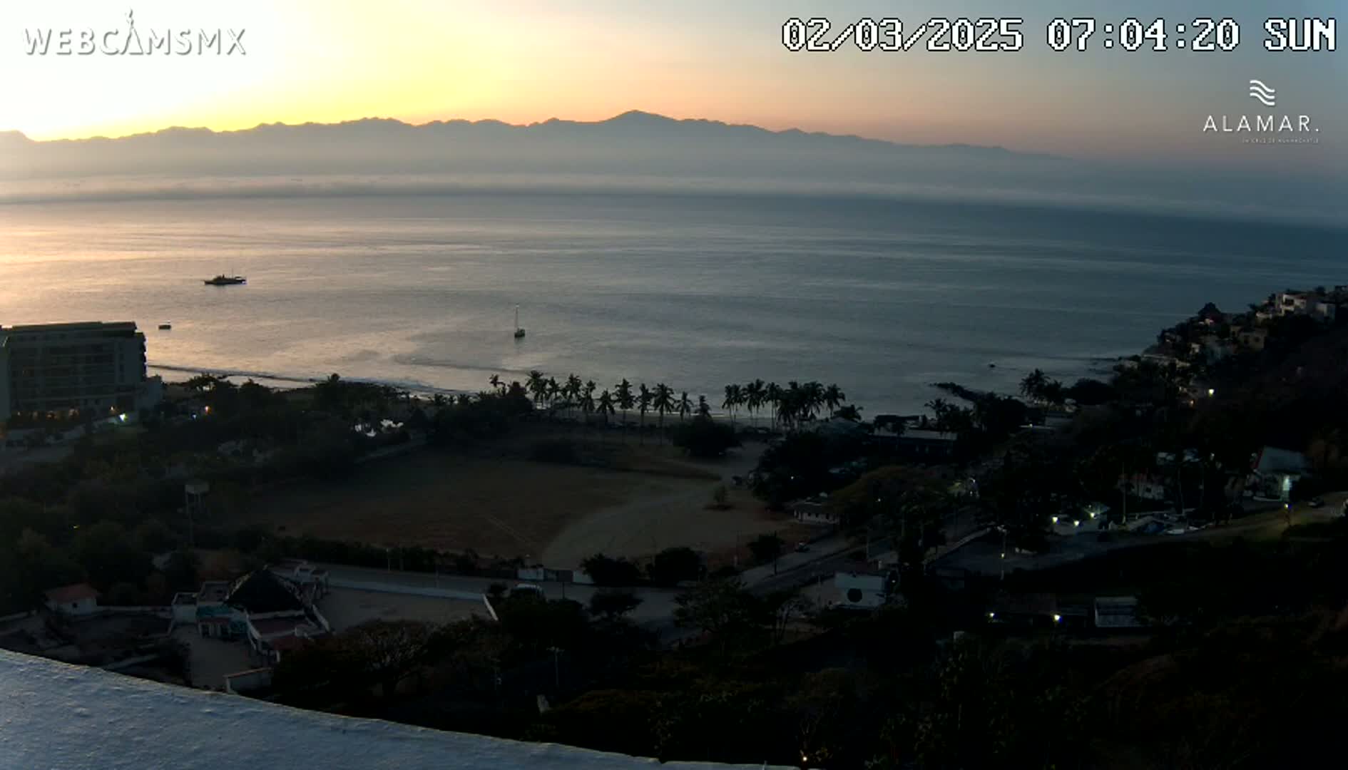 A coastal scene at sunrise shows a calm ocean, a beach with palm trees, and buildings nestled along the shoreline, with mountains visible in the distance.