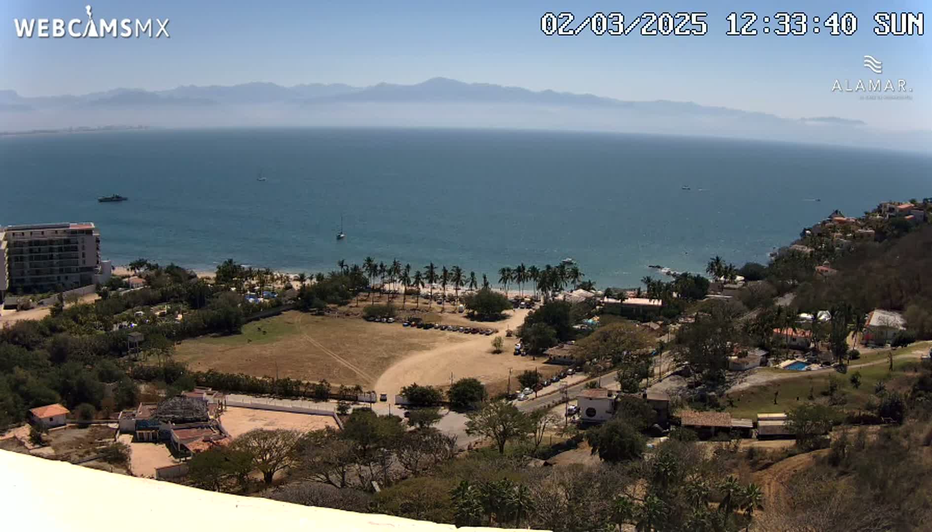 A sunny coastal view shows a calm ocean, a sandy beach lined with palm trees, a nearby town, and mountains in the distance.