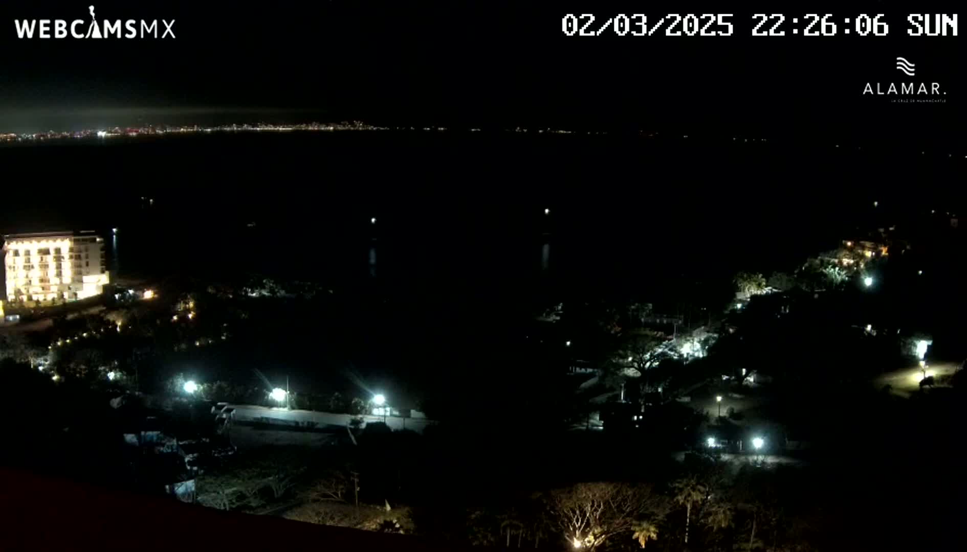 A nighttime view of a coastal area shows a building illuminated near the foreground with a dark body of water and a distant city skyline visible under a clear sky.