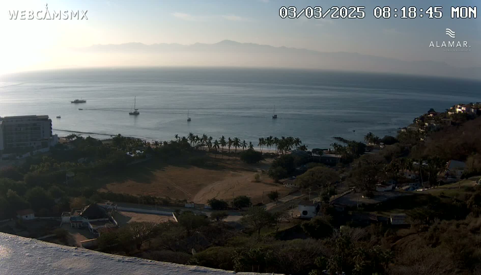 A hazy, sunny coastal view shows a calm ocean with several boats, a sandy beach lined with palm trees, and a hillside town nestled amongst dark green vegetation.