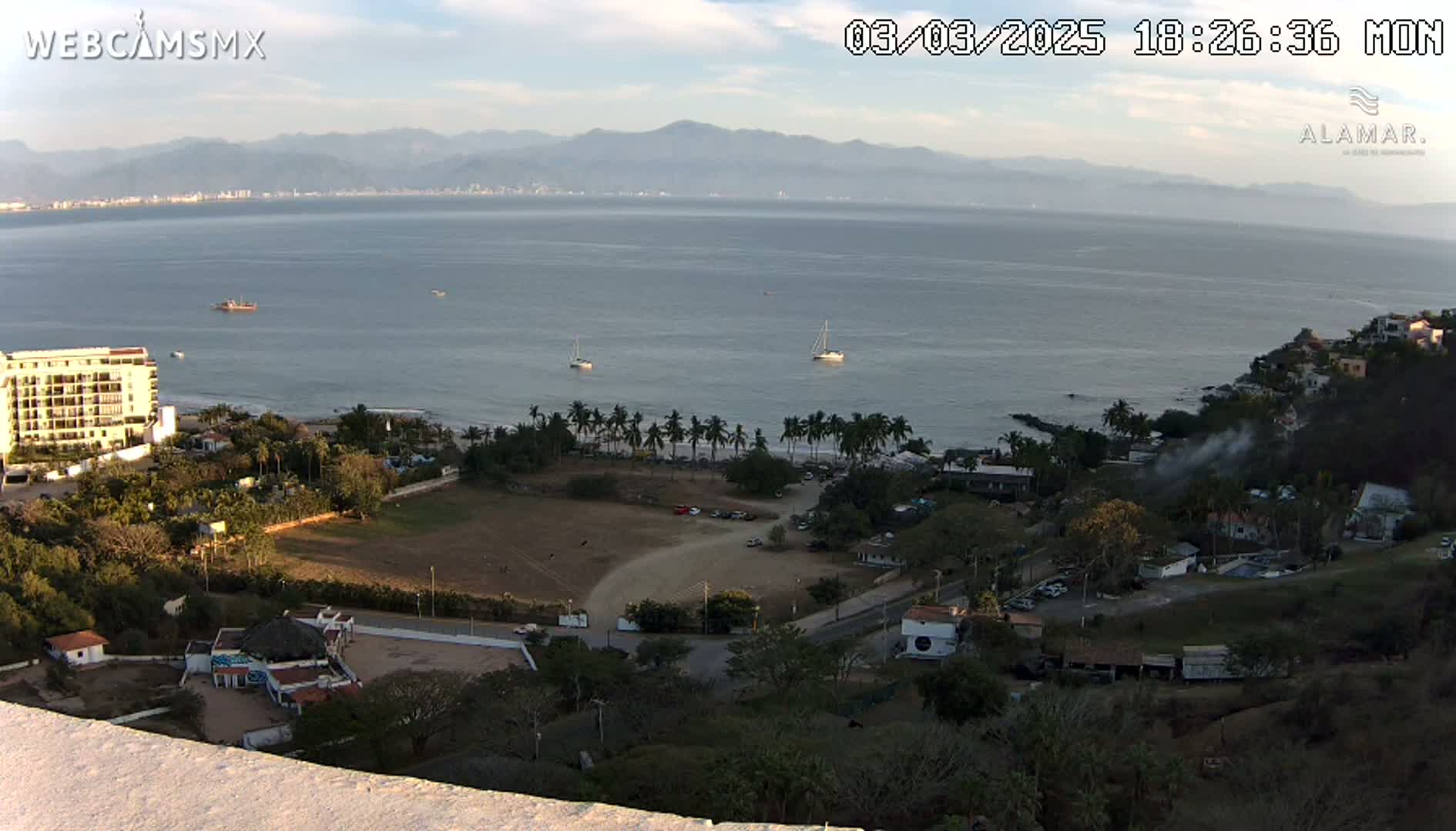 A high-angle, long shot shows a coastal town nestled beside a calm bay under a mostly clear sky, with mountains in the distance.
