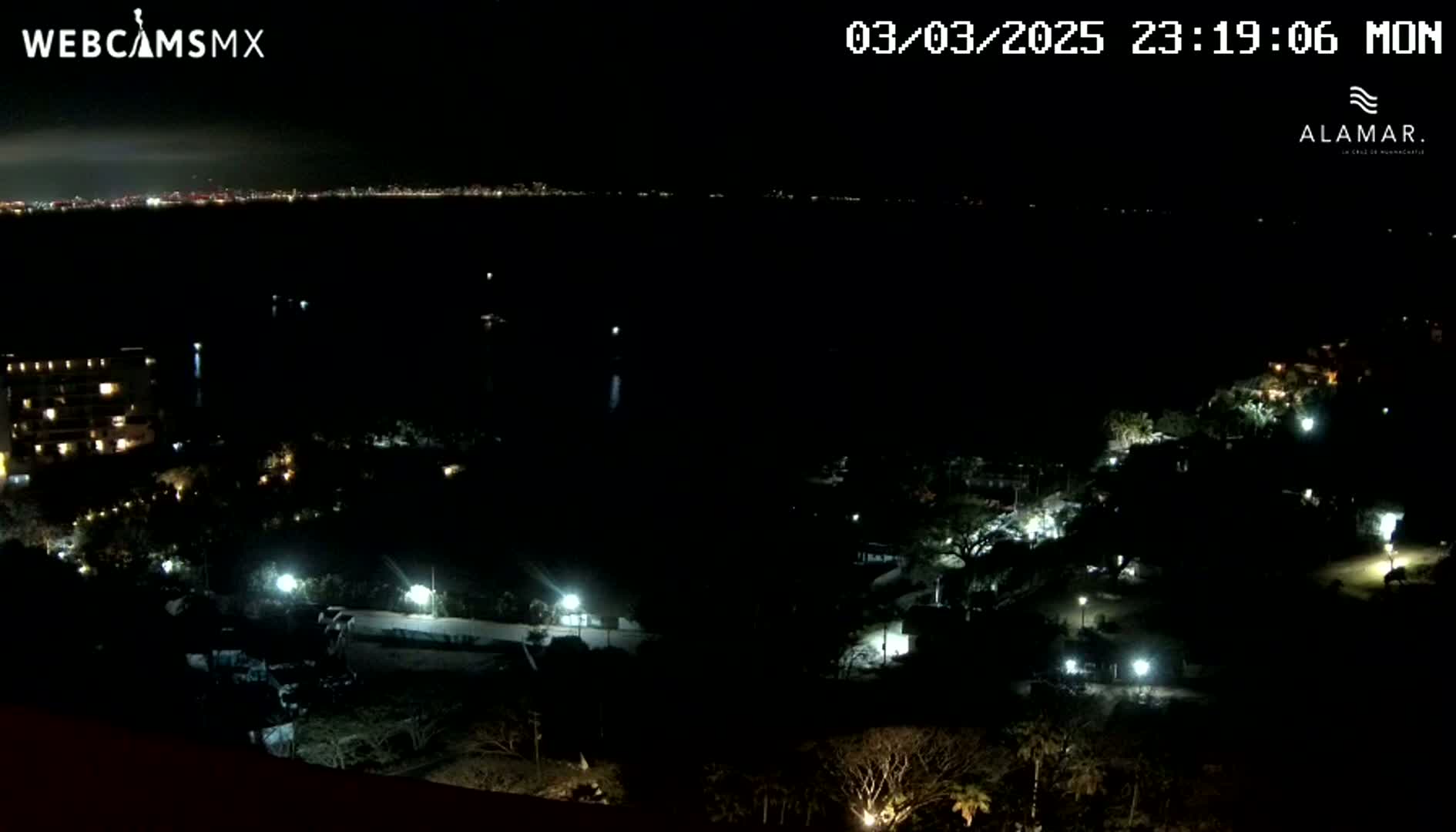 A nighttime, high-angle view shows a dark coastline with scattered lights from buildings and streetlights, overlooking a body of dark water with a distant, faintly lit city skyline.