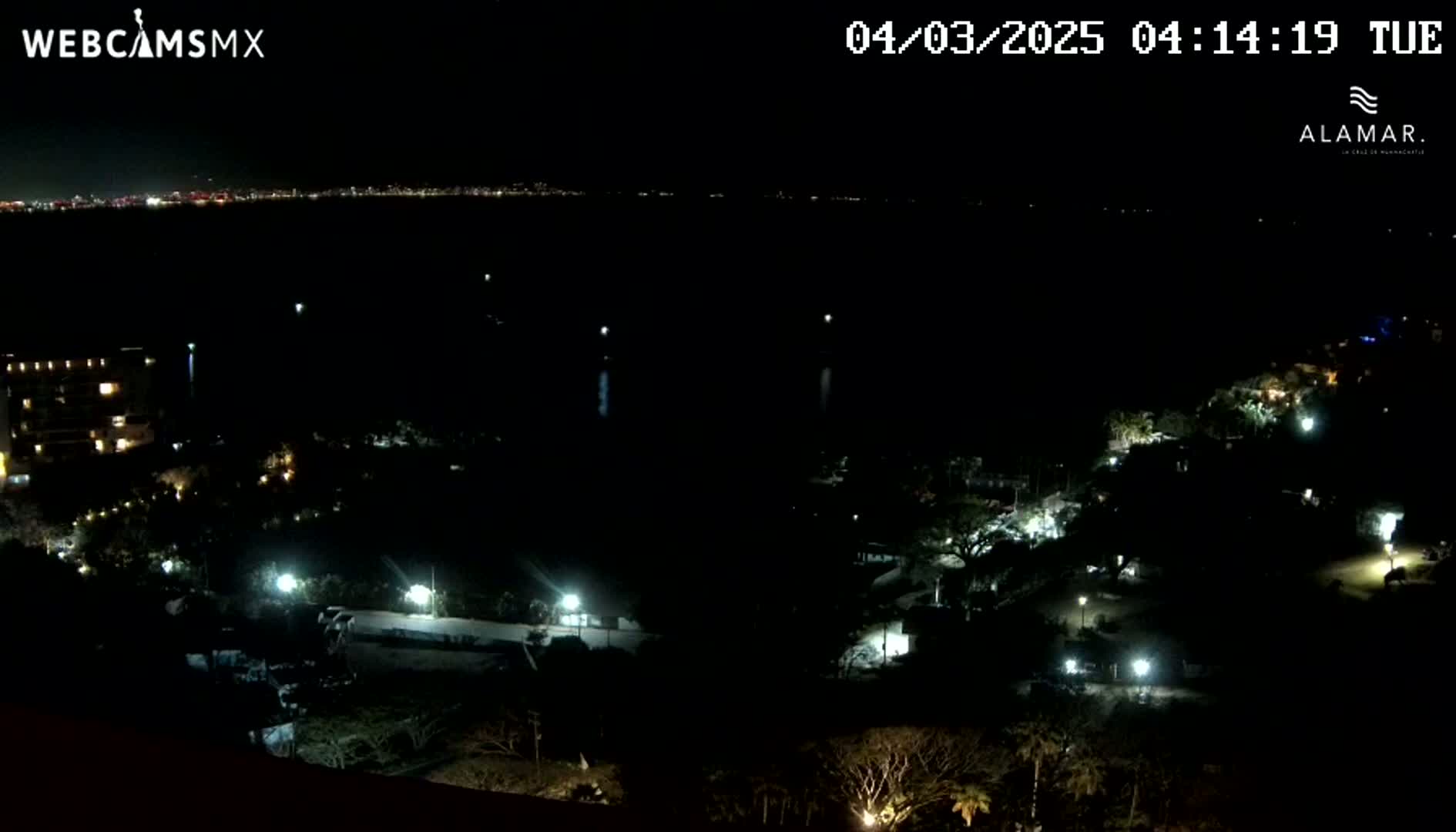 A nighttime, high-angle view shows a dark coastline with a few lights visible along the shore and in the distance, and a dark body of water with a distant city skyline illuminated.
