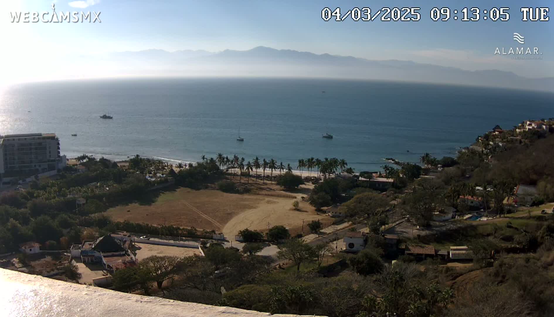 A high-angle, long shot on a sunny day shows a coastal town nestled below a hillside overlooking a calm ocean and distant mountains.
