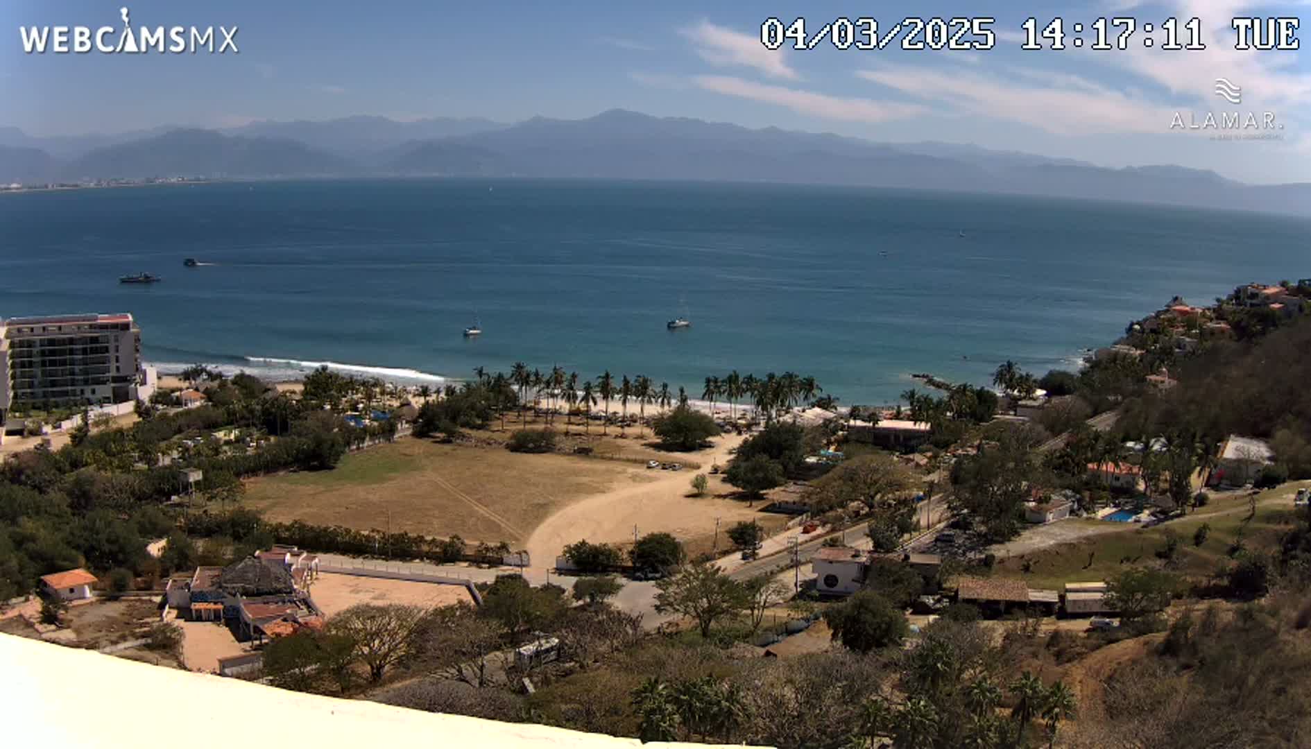 An elevated view shows a beach with palm trees, a coastline with buildings, and a calm ocean under a clear blue sky.