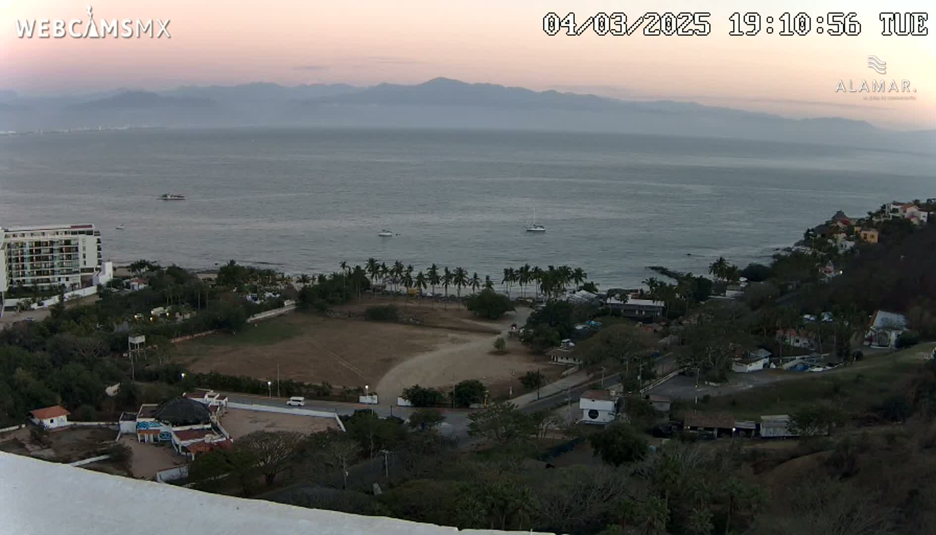 An elevated view shows a coastal town at dusk, with a calm ocean extending to distant mountains under a clear, twilight sky.