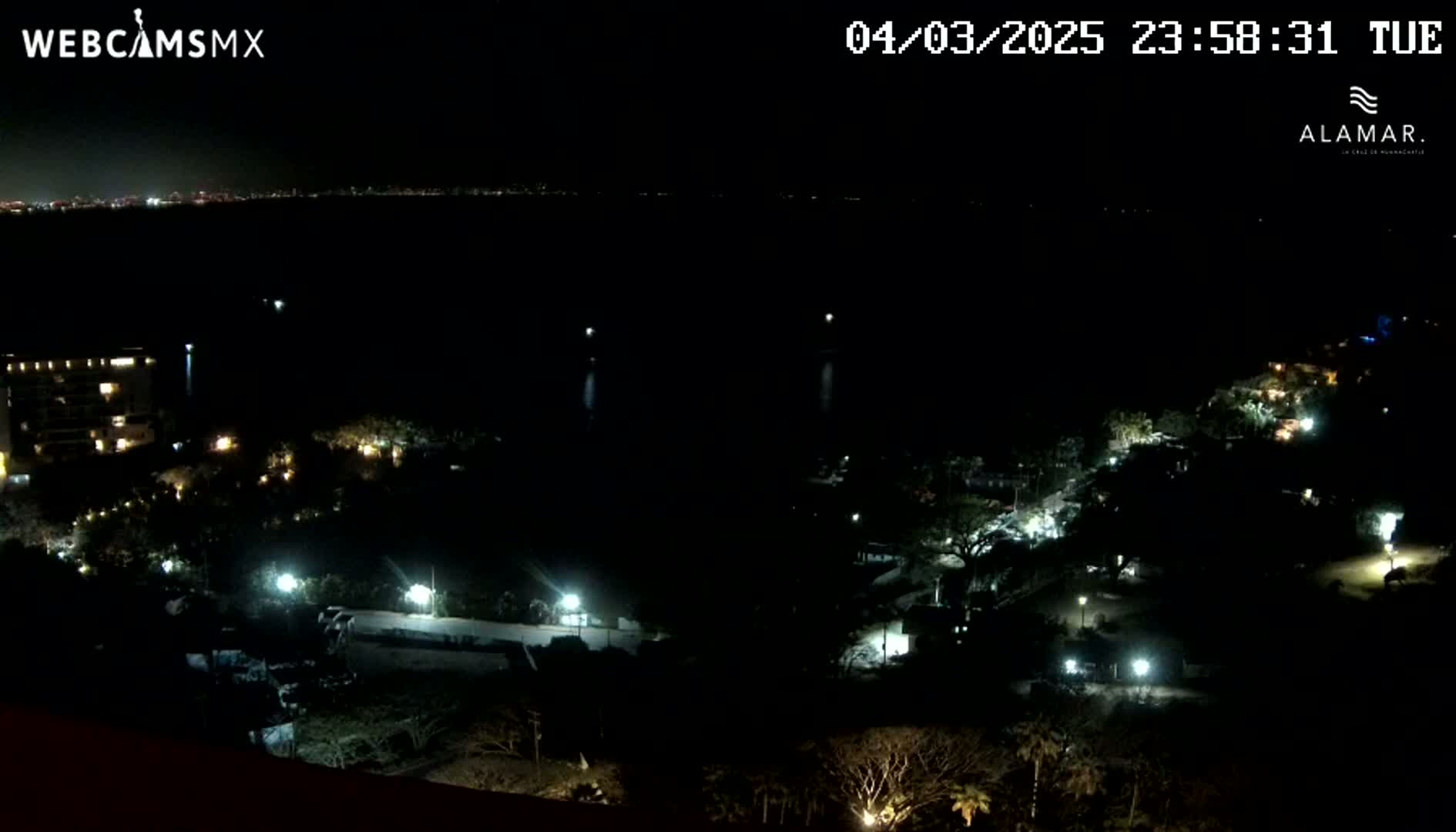 A nighttime, high-angle view of a coastal area shows a dark body of water, sparsely lit buildings and trees along the shore, and a faintly visible cityscape in the distance.