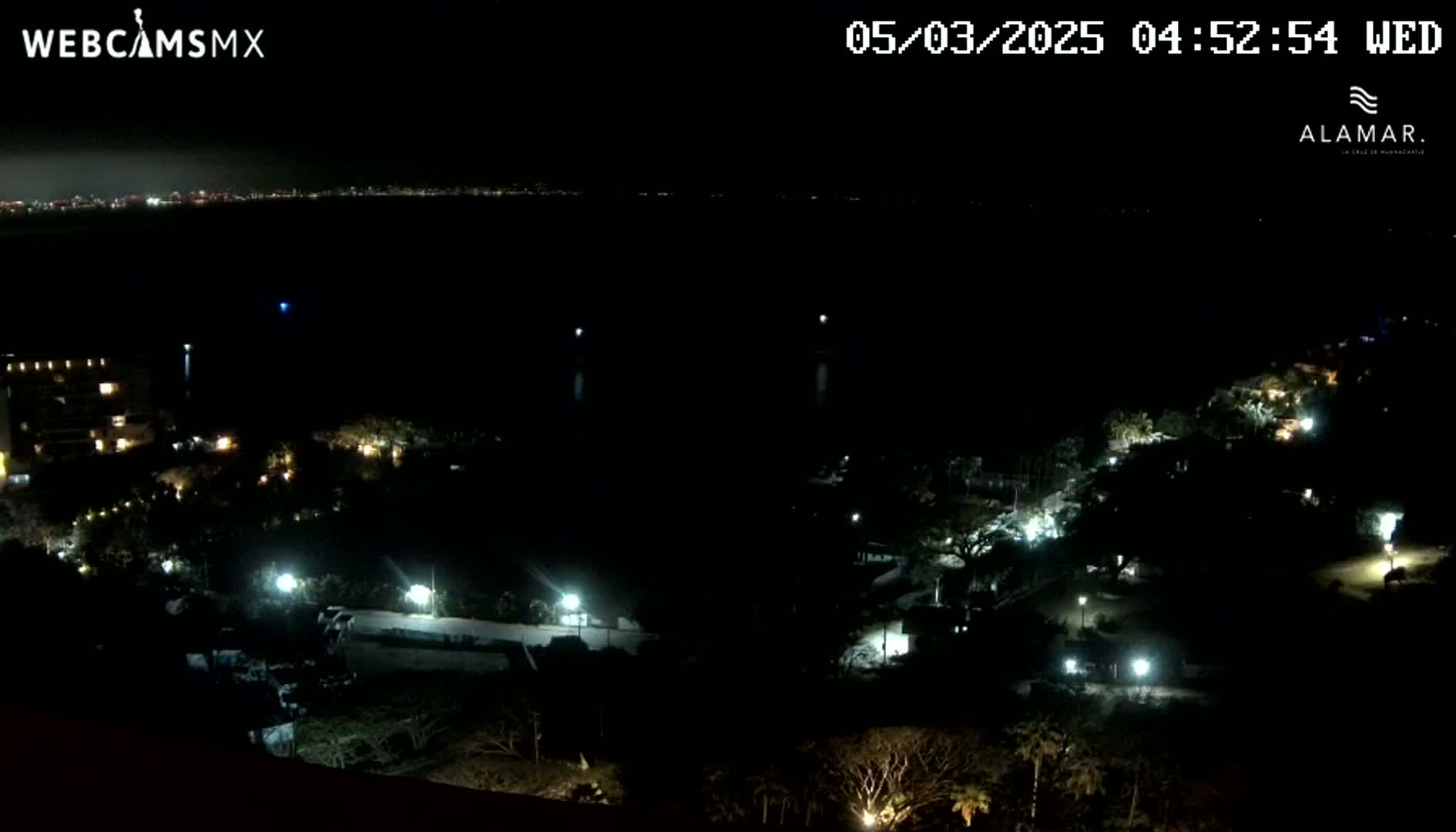 A nighttime, long-range view shows a dark body of water with a distant city skyline, and a hill illuminated by scattered lights from buildings and street lamps.
