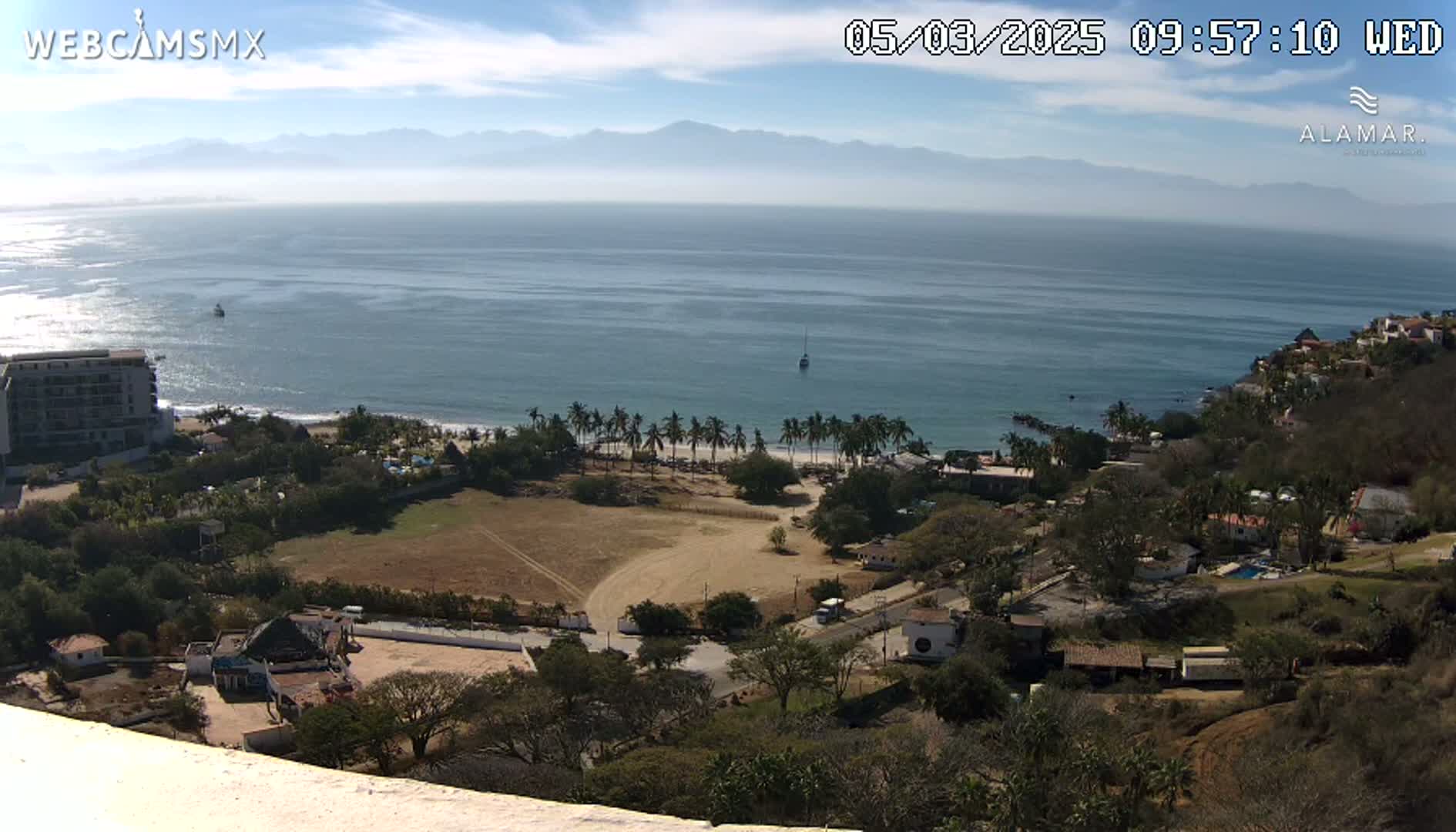 An elevated view shows a coastline with a calm ocean under a partly sunny sky, overlooking a beach with palm trees, buildings, and a sandy area.
