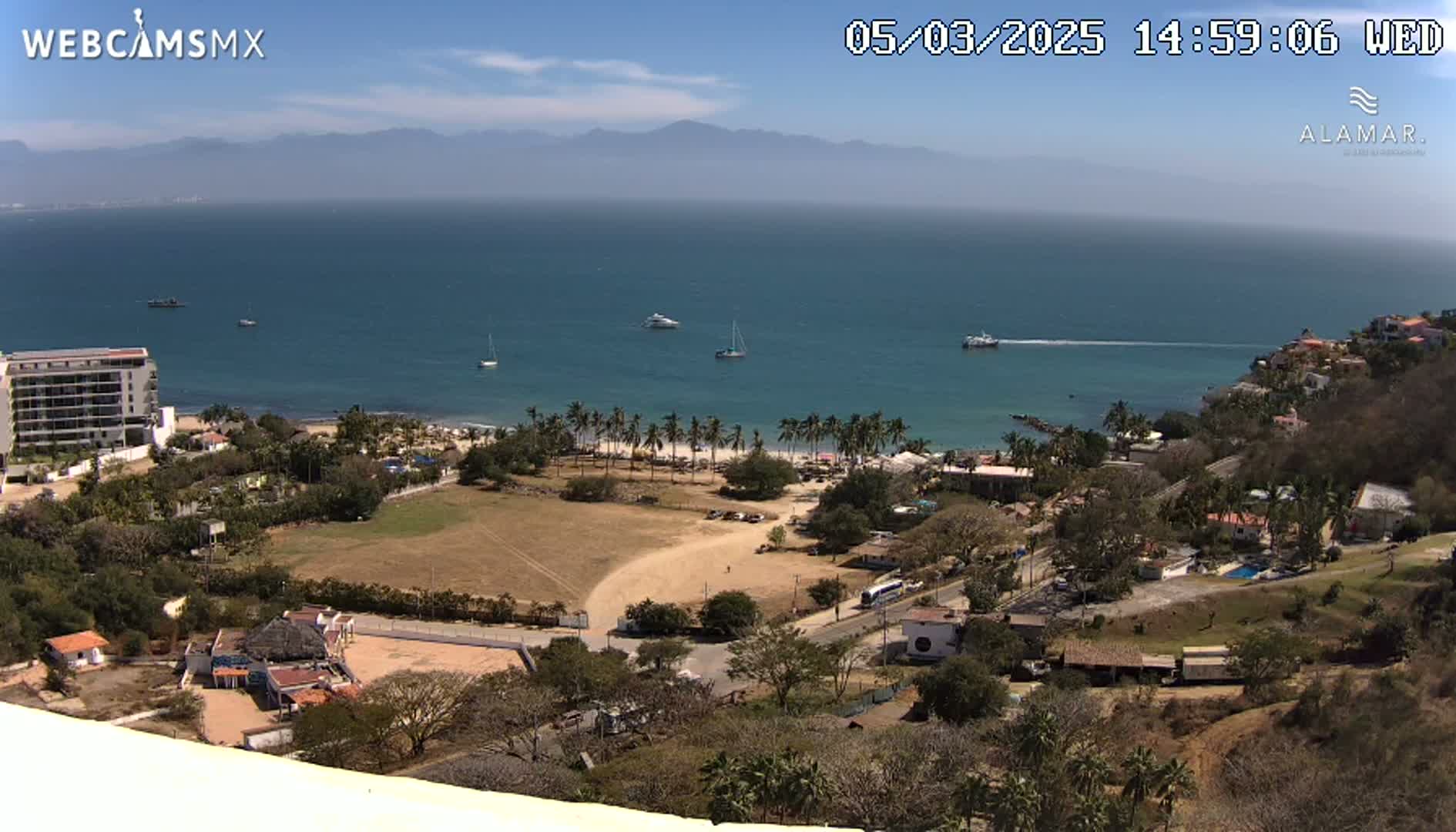A high-angle, sunny view of a coastal town with a sandy beach, palm trees, several buildings, and boats on a calm ocean, mountains visible in the distance.