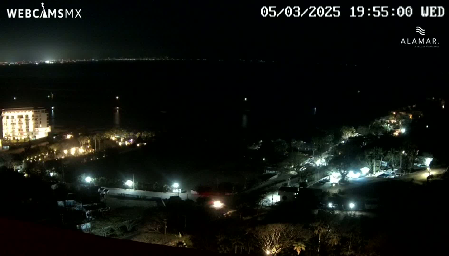 A nighttime coastal scene shows a dark body of water, a brightly lit building near the water, and other illuminated areas of land, all under a clear, dark sky.