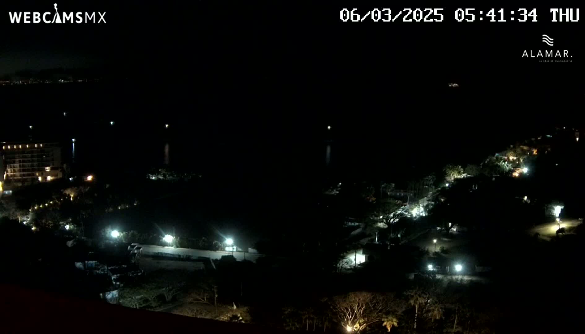 A nighttime, high-angle view shows a dark body of water, a shoreline with sparsely lit buildings and vegetation, and a distant, dark cityscape under a clear sky.