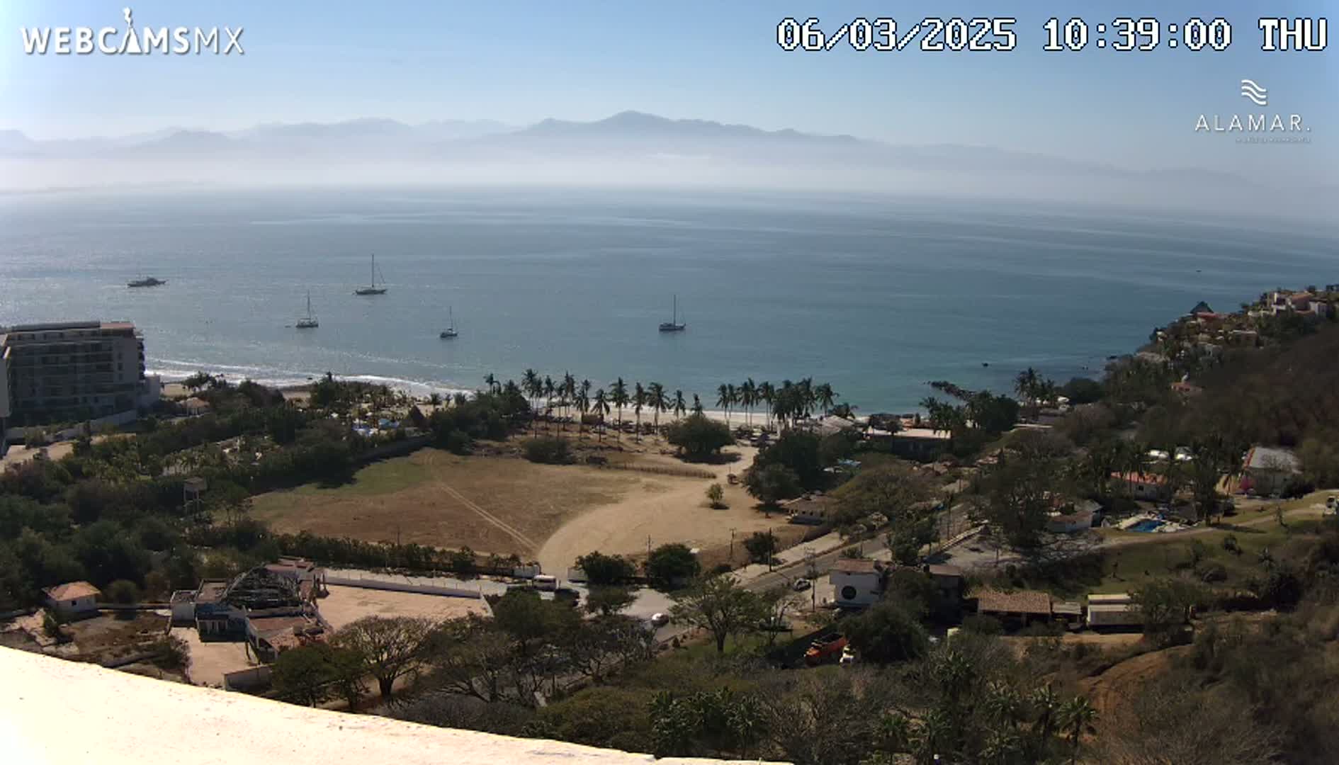 An elevated view shows a coastline with a sandy beach lined with palm trees, several boats in the ocean, and buildings nestled among vegetation under a clear, sunny sky.