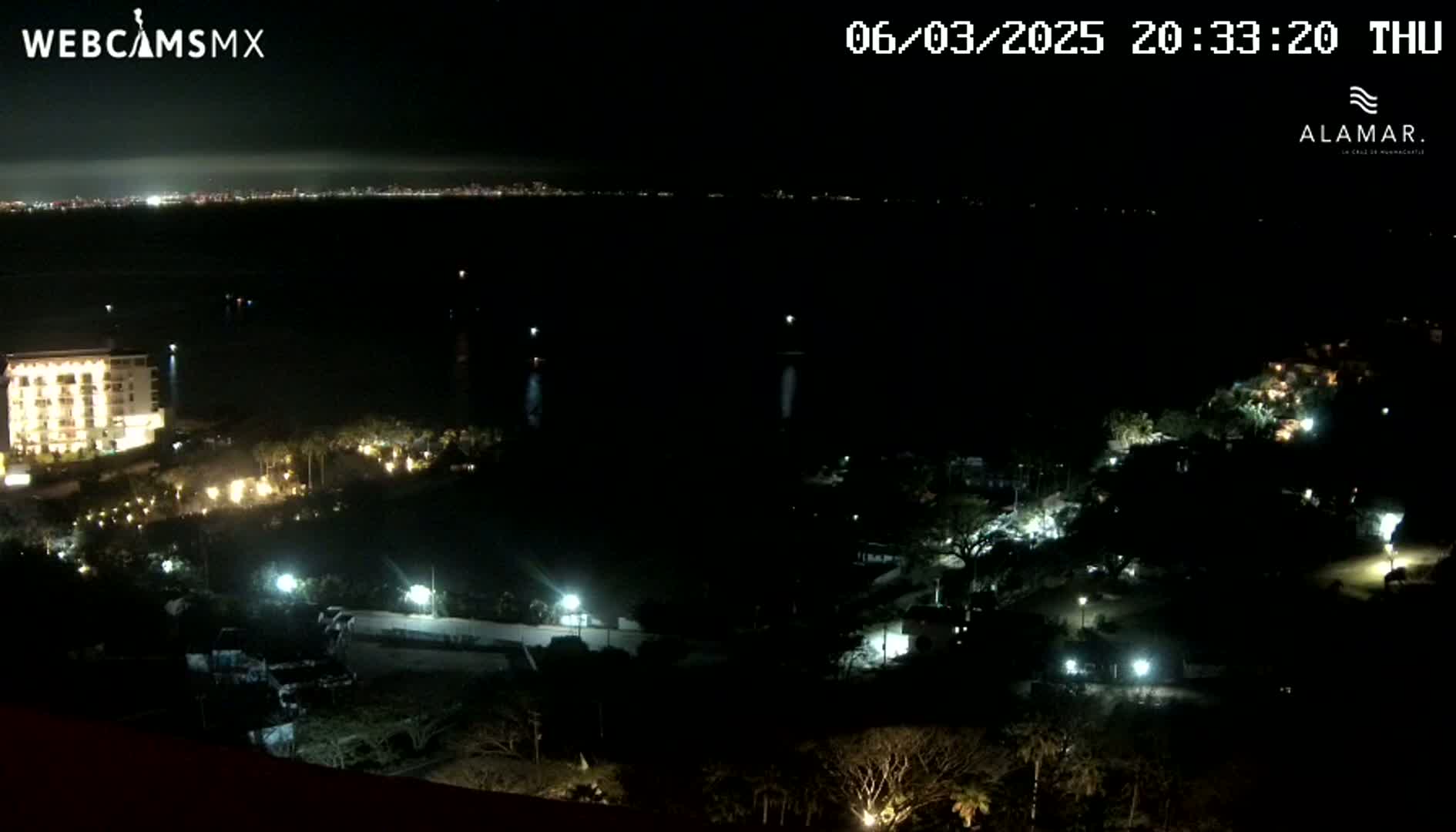 A nighttime view shows a dark body of water with a city skyline faintly visible in the distance, along with a lit-up hotel and surrounding grounds closer to the viewer.
