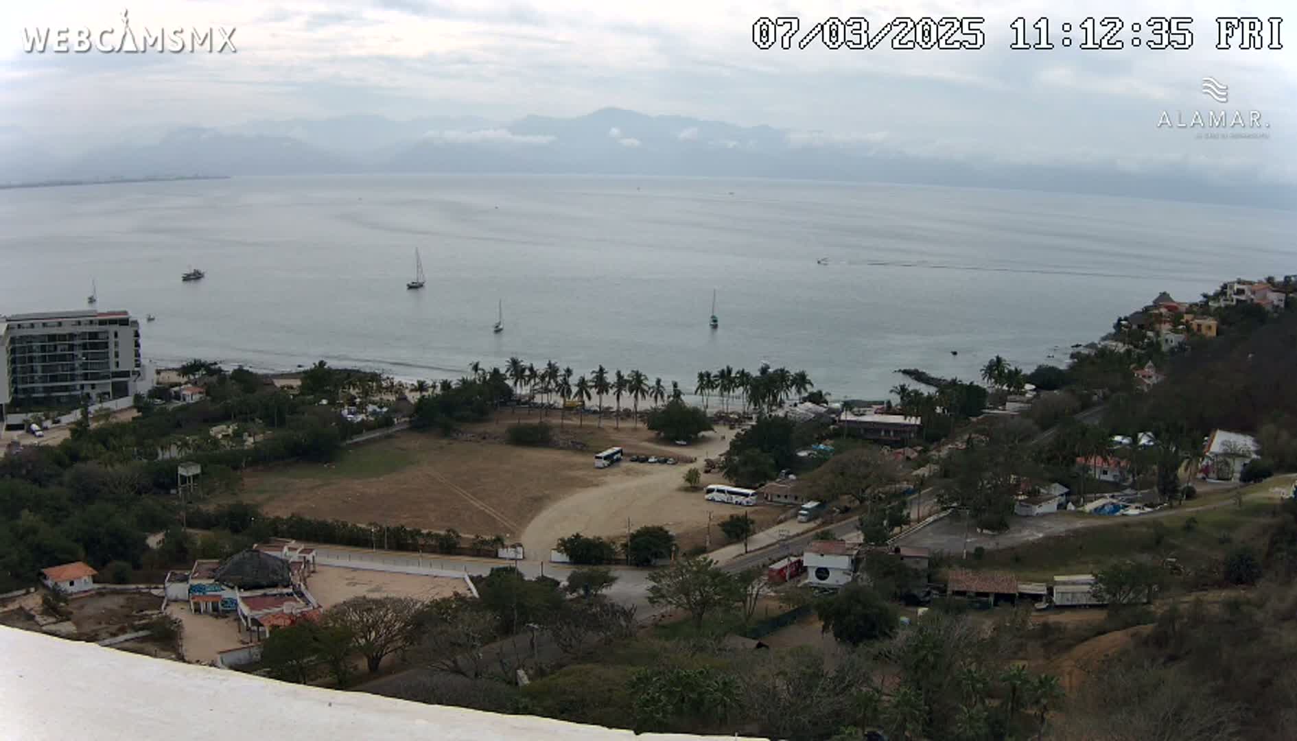 A high-angle view of a coastal town on a partly cloudy day, showing a sandy beach, calm ocean water with several boats, a parking lot with buses, buildings, and lush vegetation on a hillside.